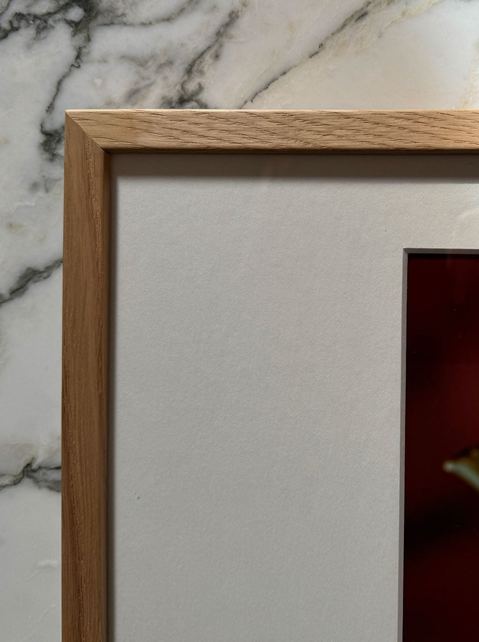 A close-up of a wooden picture frame displaying a fine art print. The frame is light wood with a matte white border surrounding the image. The background features a marble surface with grey and white veining, and only a corner of the limited edition artwork "Apple and Pearls" by Adeline Mai is visible.