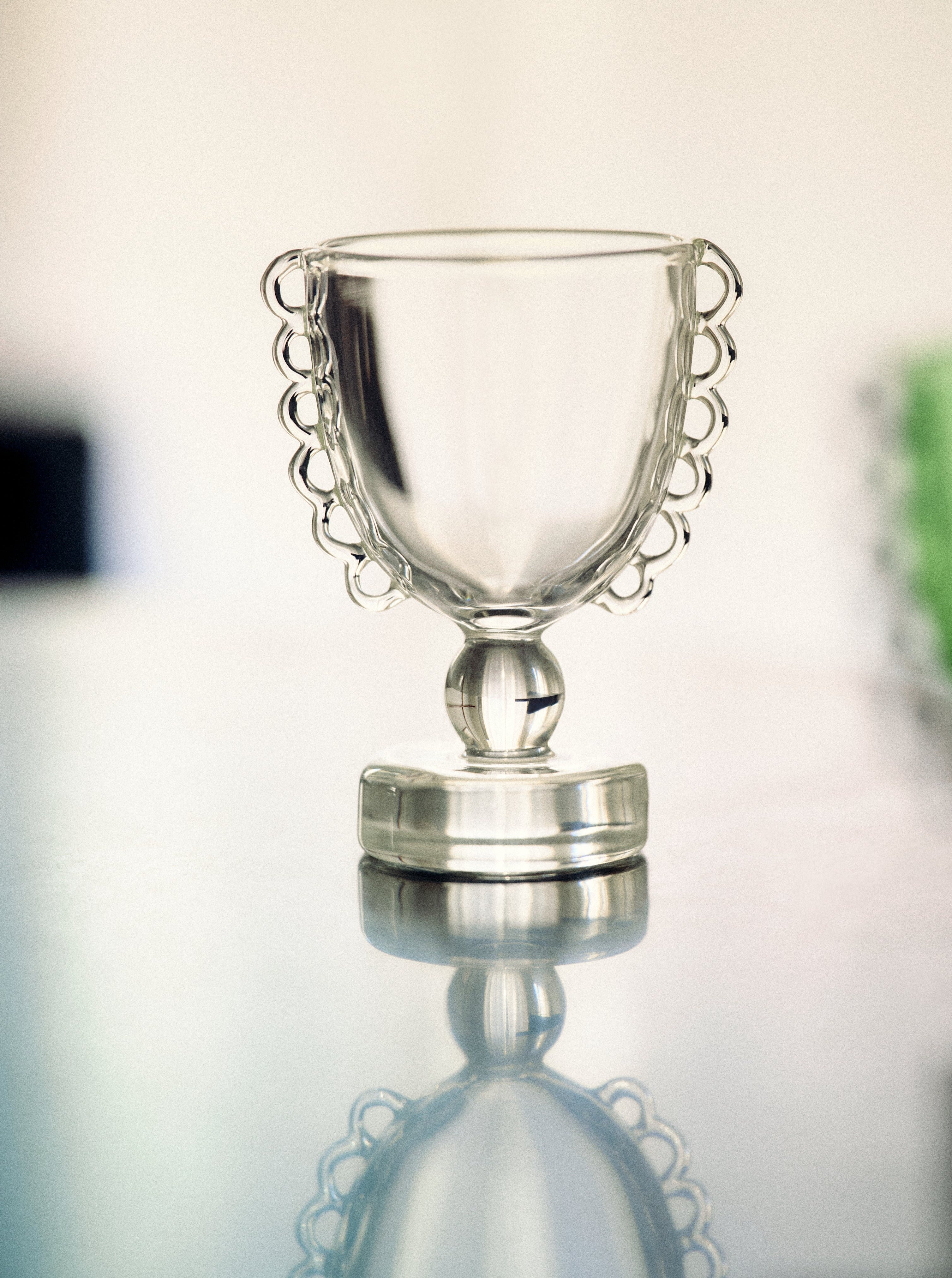 A small, intricately designed Coco Egg Cup Set by Sophie Lou Jacobsen, reminiscent of a vintage salt dish, with handles on either side is placed on a reflective surface. The background is blurred, focusing attention on the delicate details of the egg cup set.