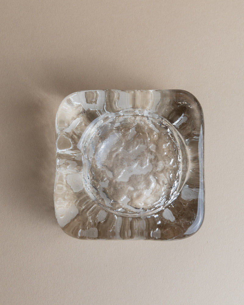 Resting on a beige surface, the Vintage Murano Glass Ashtray by Bottega Jacobs features slightly rounded edges and a textured pattern that plays with reflections, exuding retro charm. Its translucent glass shows interior wear, enhancing its vintage appeal.