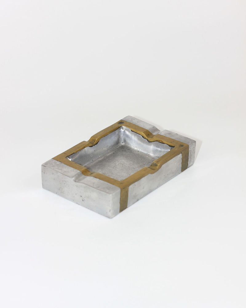 The Brutalist Ashtray 70s by Boga Avante Shop has a rectangular silver body with gold trim and side grooves for cigarettes, made in Spain. Seen on a plain white background.