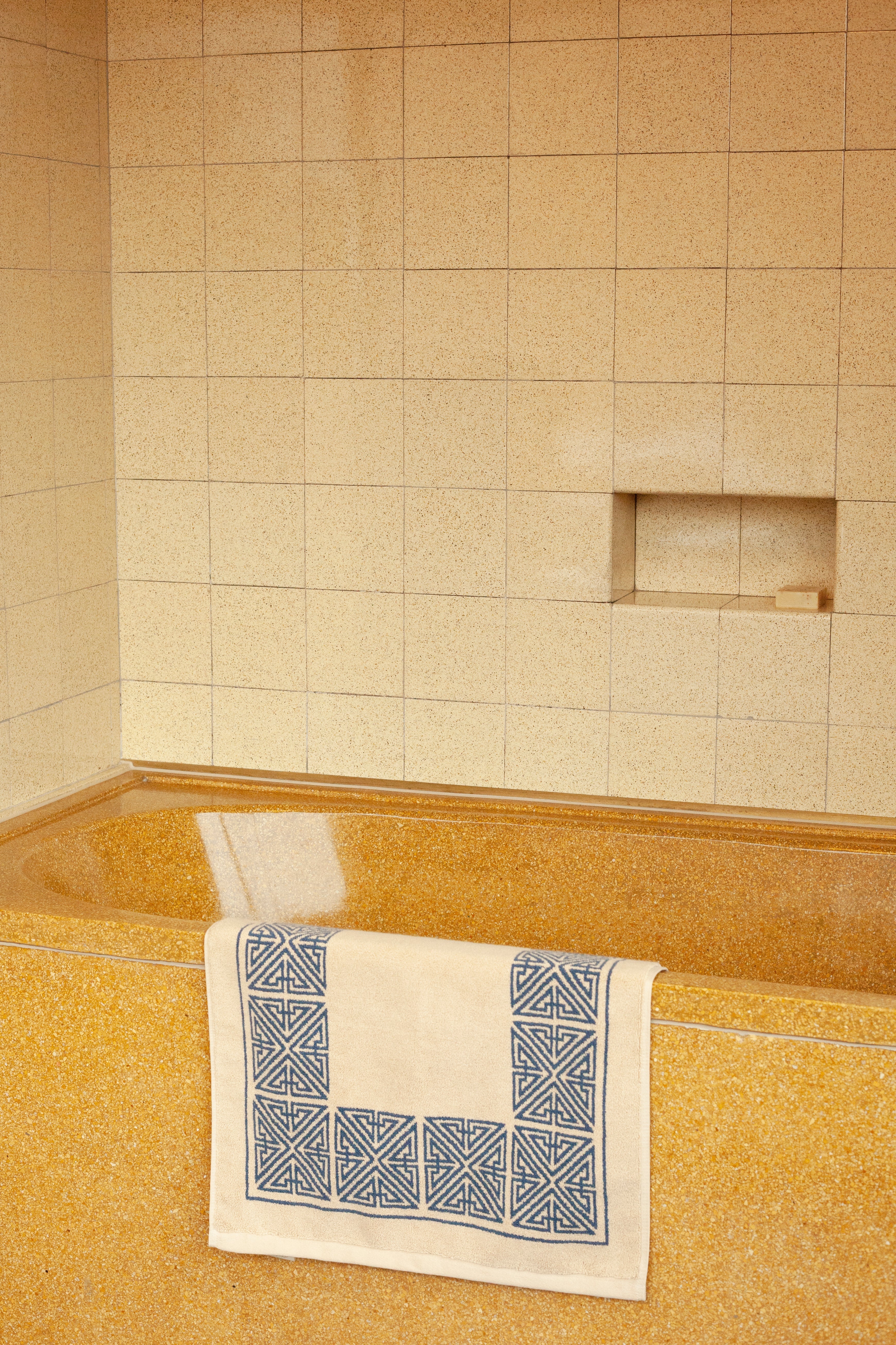 A minimalist bathroom corner with textured yellow wall tiles and a matching yellow floor now features an Agnes bath mat in marine blue by Autumn Sonata, placed on the floor.