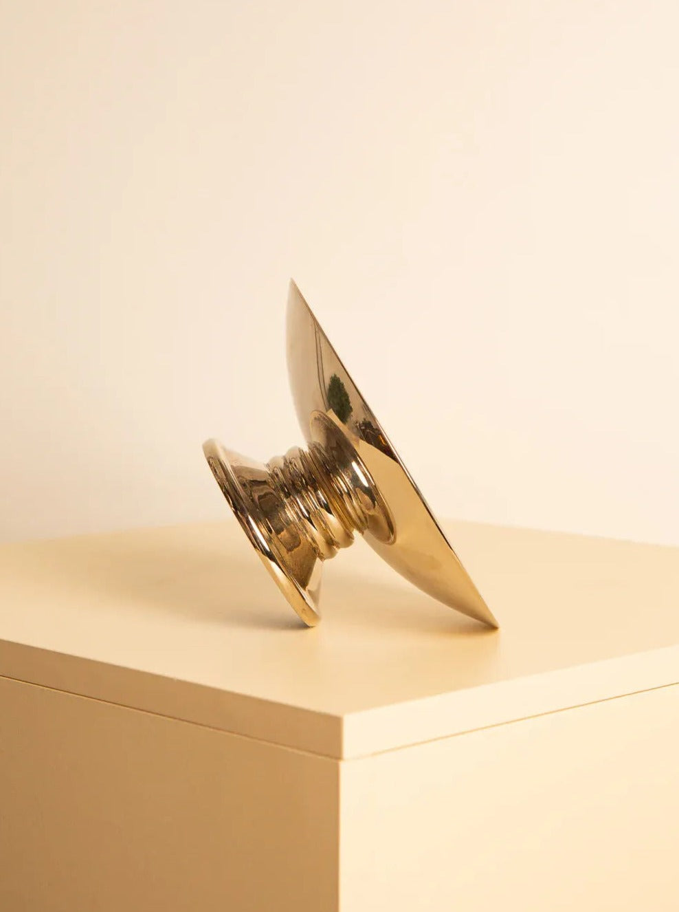 A shiny, Treaptyque 80's Silver Plated Bowl resembling a wide, shallow dish with a conical base is placed on its side atop a beige rectangular surface. The object contrasts with the off-white wall in the background, highlighting its impeccable condition.