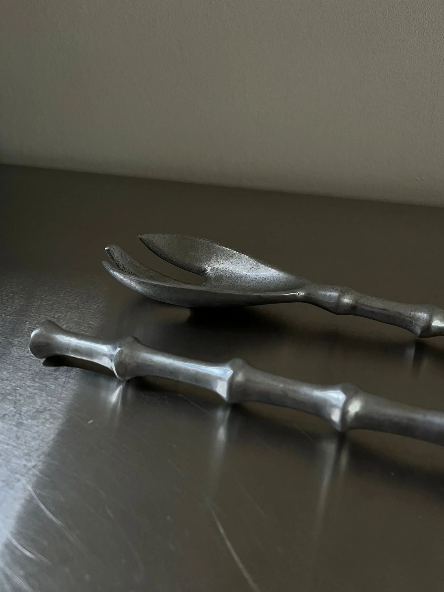 A close-up image of two silver-plated utensils with anthropomorphic handles. One utensil resembles a fork with two prongs, while the other appears to be a spoon. They are placed on a metallic surface against a plain wall. The handles have a textured, irregular design. The utensils are part of the Lettuce Cutlery set by CU-RA-TED.