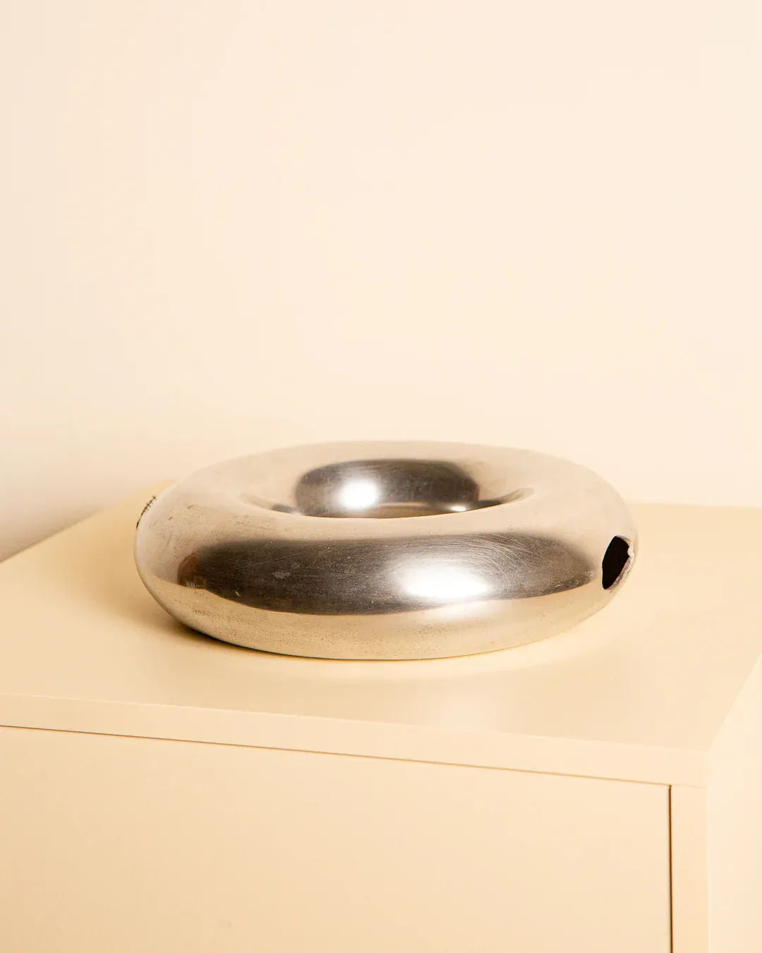 A round, shiny aluminum sculpture, the 80's Aluminum "Circle" Vase by Treaptyque, resembling a donut, sits on a light-colored table against a cream background, echoing an Italian style similar to the Cercle vase.