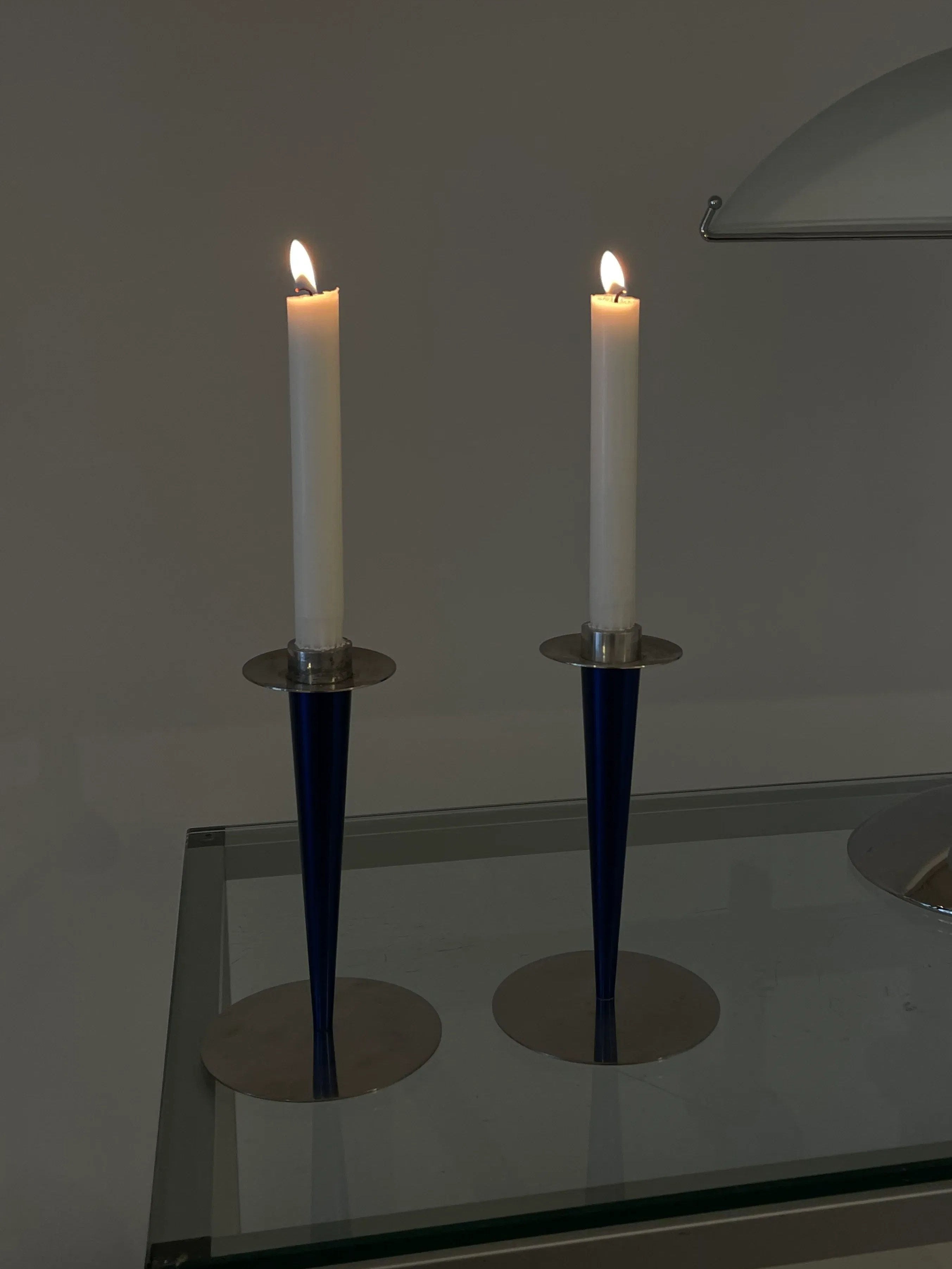 Two white candles are lit and placed in Collect Cph's Vintage Steel Candle Holders with circular stainless steel bases. The setup is on a glass surface against a grayish background, with the glow from the candles providing a warm ambiance.