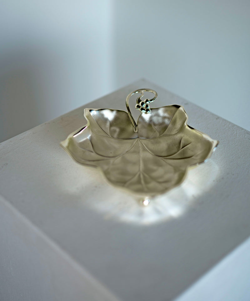 A Grape Leaf Dish by Porter Studio, showcasing intricate vein details reminiscent of the 1930s, is elegantly displayed on a white square pedestal against a plain background. Its soft shadows and timeless design evoke an era of refined taste.