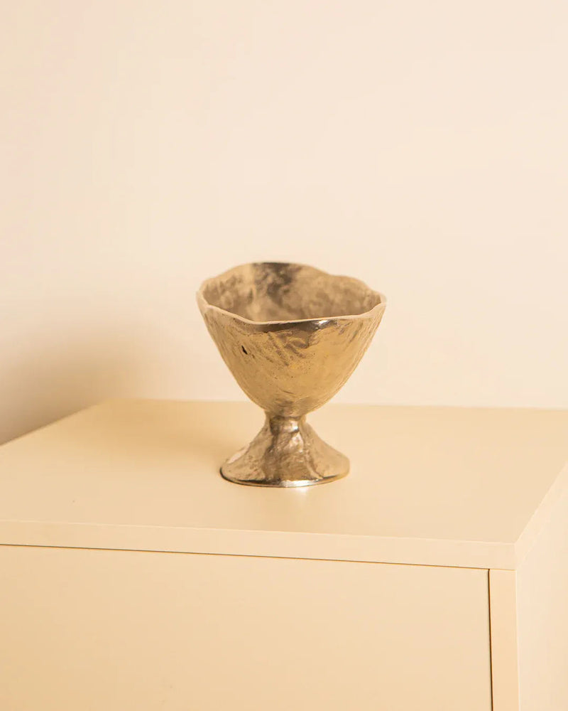 The Treaptyque Large Italian Bowl in Cast Aluminium 80's, with its gold texture and small pedestal base, sits on a light beige surface against a soft off-white background, exuding minimalistic elegance inspired by 1980s Milan design.