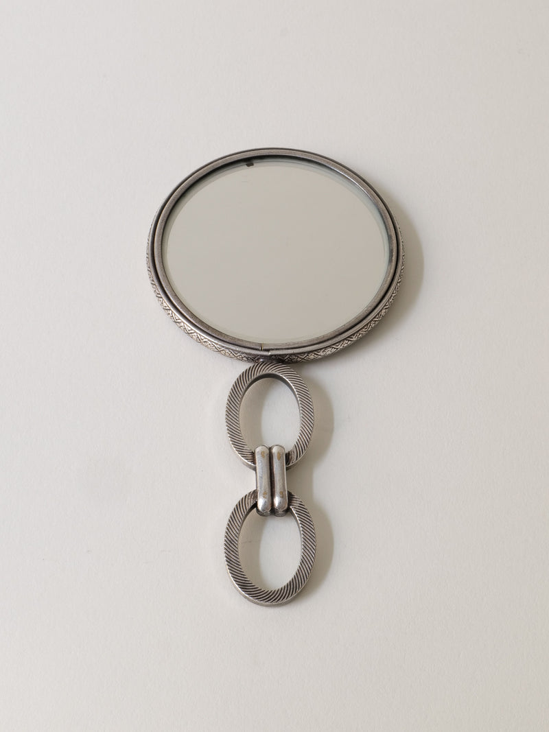 The "Hand Mirror Jean Després 1920s" by Collection apart features a round mirror with a decorative metallic handle, showcasing two interlocking rings with a textured design, evoking Art Deco flair inspired by Jean Després.