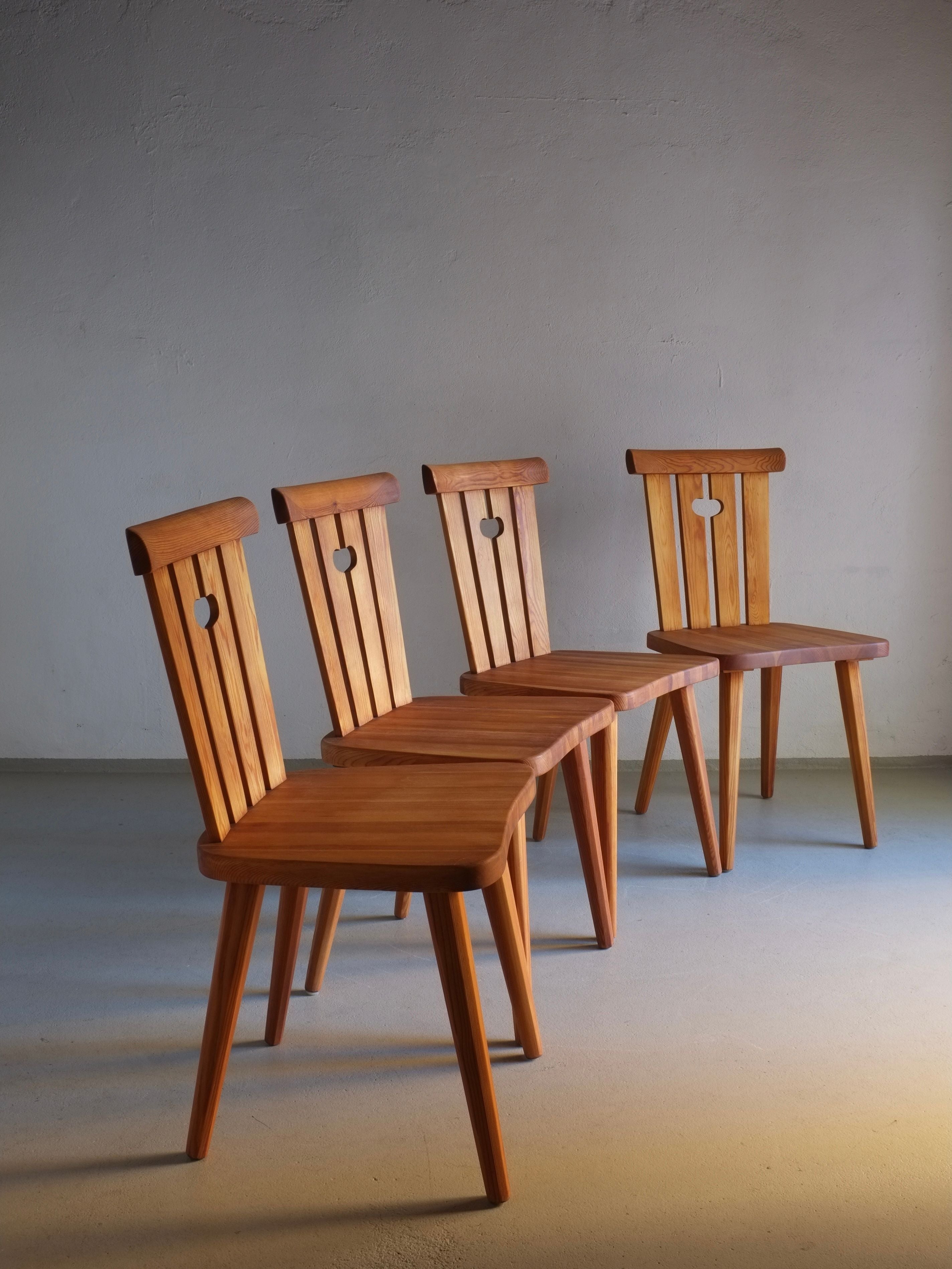 Four rustic pine chairs by Goran Malmvall from the 1940s, restored and oiled, feature heart-shaped cutouts in their backs and are neatly lined up on a smooth, light-colored floor against a plain wall. The warm tones of the wood from these Veter Vintage pieces add a cozy feeling to the simple, minimalist setting.