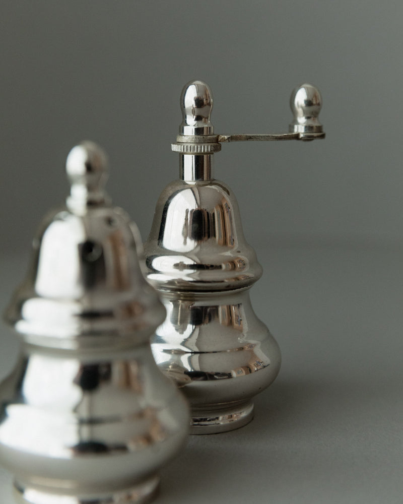 Close-up of Bottega Jacobs' Salt Shaker and Pepper Set - Italy 1970s on a neutral background. The vintage-style polished silver-plated mills feature a classic reflective design, with one showcasing a small crank handle on top.