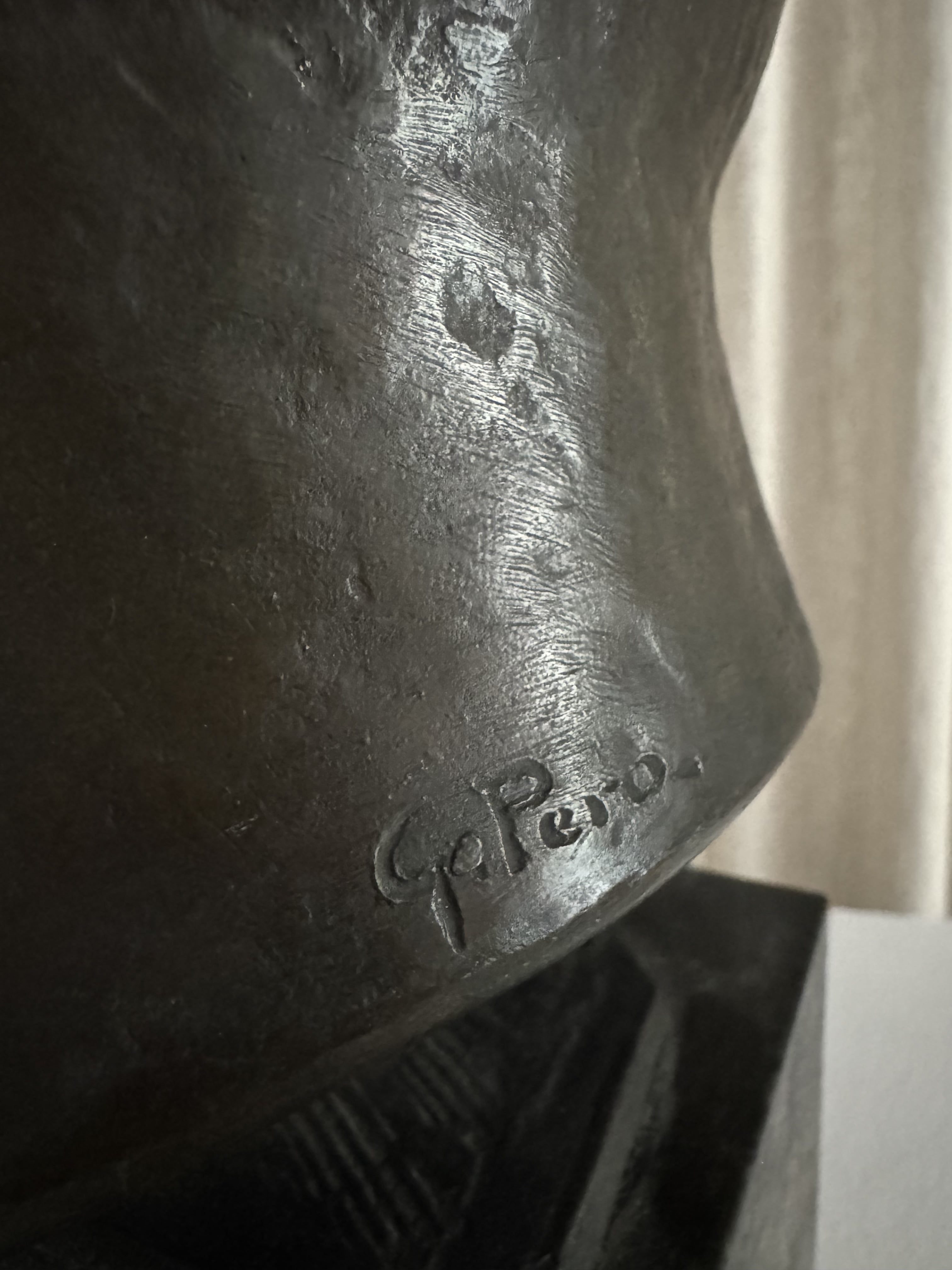 Close-up of FEBRERO SHOP's bronze sculpture, TESTA VIRILE, highlighting the signature engraved on its surface. The texture of the metal and detailed craftsmanship stand out against a light-colored background.