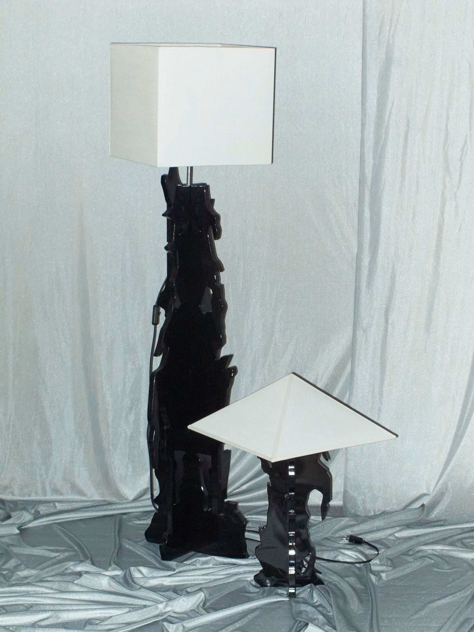 Two contemporary lamps with artistic bases are displayed against a draped fabric backdrop, creating a conceptual piece. The taller lamp, featuring a square shade, and the shorter one, with a triangular shade, both belong to the "The Bird" collection by Ochoa Rocabert. Their sleek black bases exhibit abstract shapes that enhance their artistic appeal.