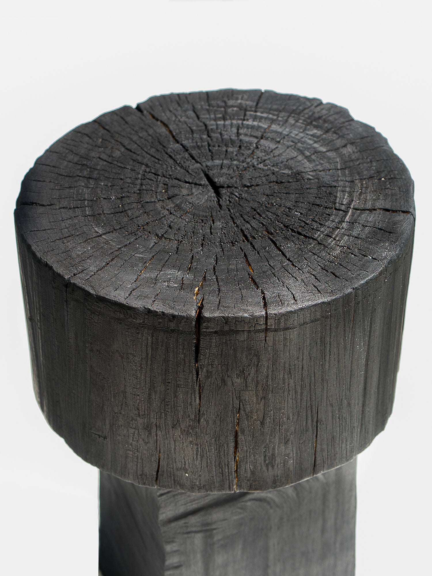 A close-up of a wooden post mirrors the artistry of Touch With Eyes' Burnt Black Coffee Tables/Stools. Its charred, cracked surface and circular grain patterns give a weathered, darkened look with radial fissures against a neutral light background.