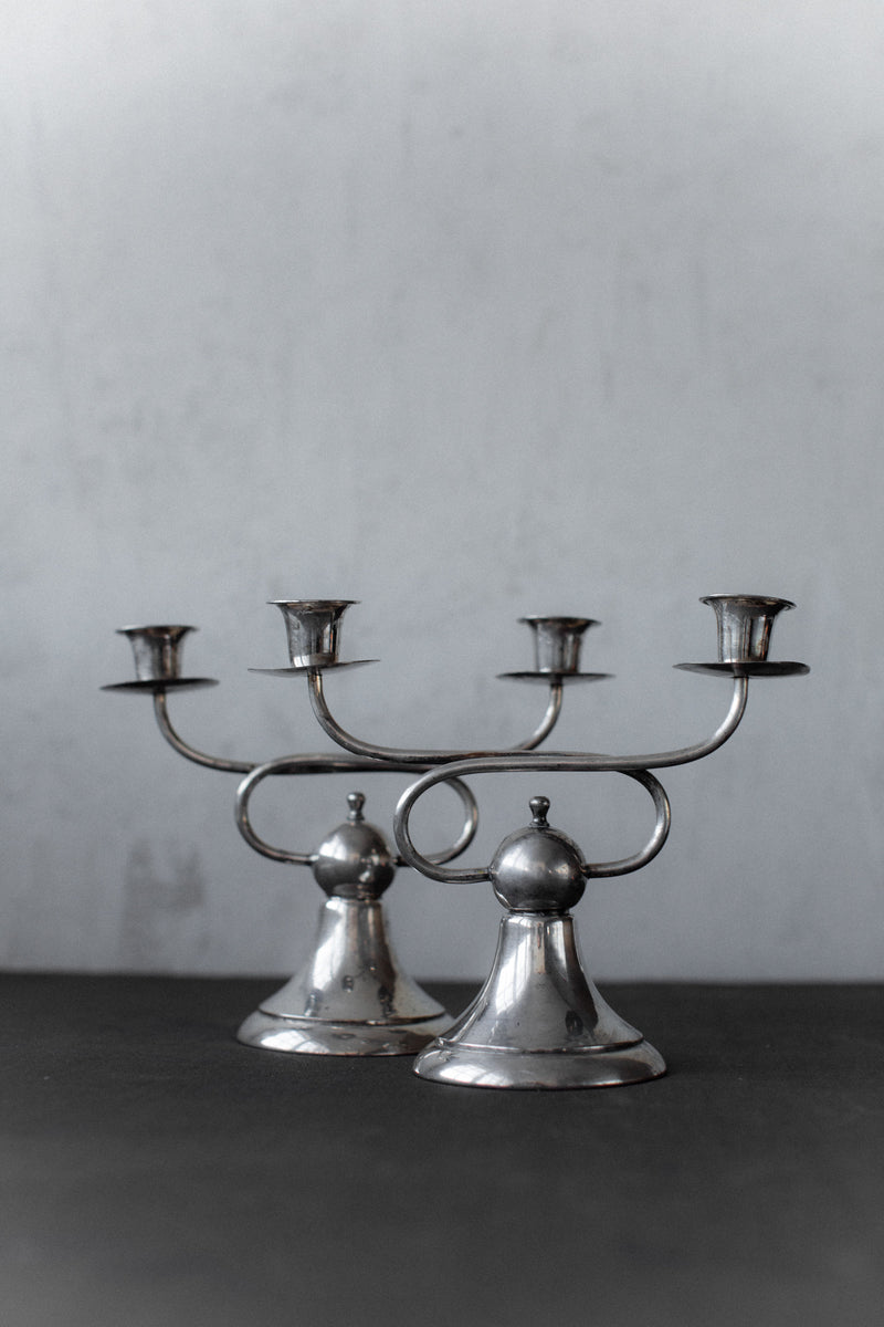 Two Art Deco Candleholders by Out For Lunch, each with three arms, sit on a dark surface against a light gray background. Their geometric elegance and minimalist lines reflect vintage Art Deco style.