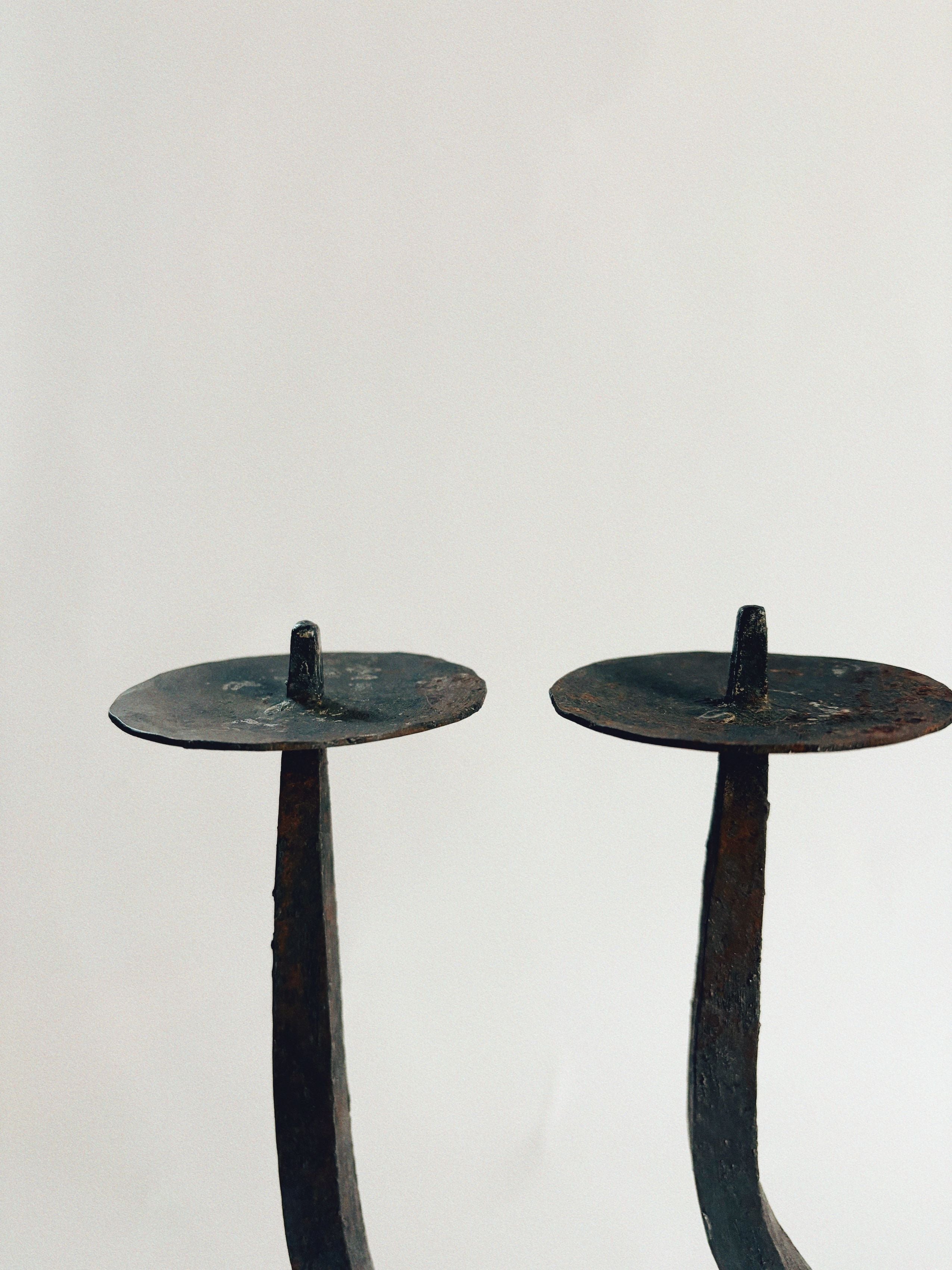 Two "Hand Forged Candleholder" by Huerto stand side by side against a plain background. These rustic, wrought iron holders feature a weathered appearance and boast a simple design with flat circular tops and slender, slightly curved stems, echoing an artisan handcrafted charm.