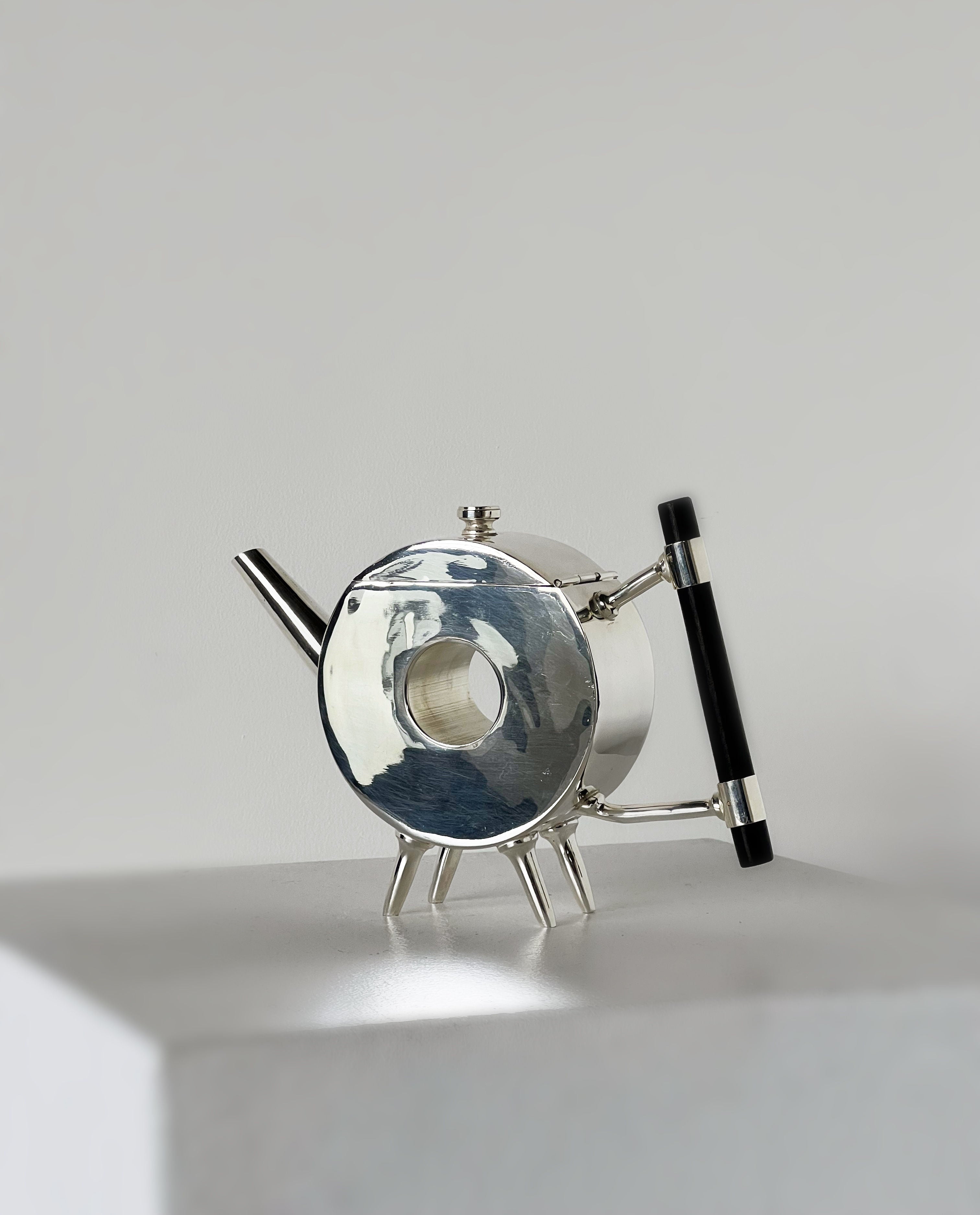 The Christopher Dresser Ornate Circular Teapot by Porter Studio, with its unique design and three pointed legs, sits on a white surface. Crafted from silver-plated metal, it features a narrow spout and a shiny reflective body that creates intriguing light patterns.