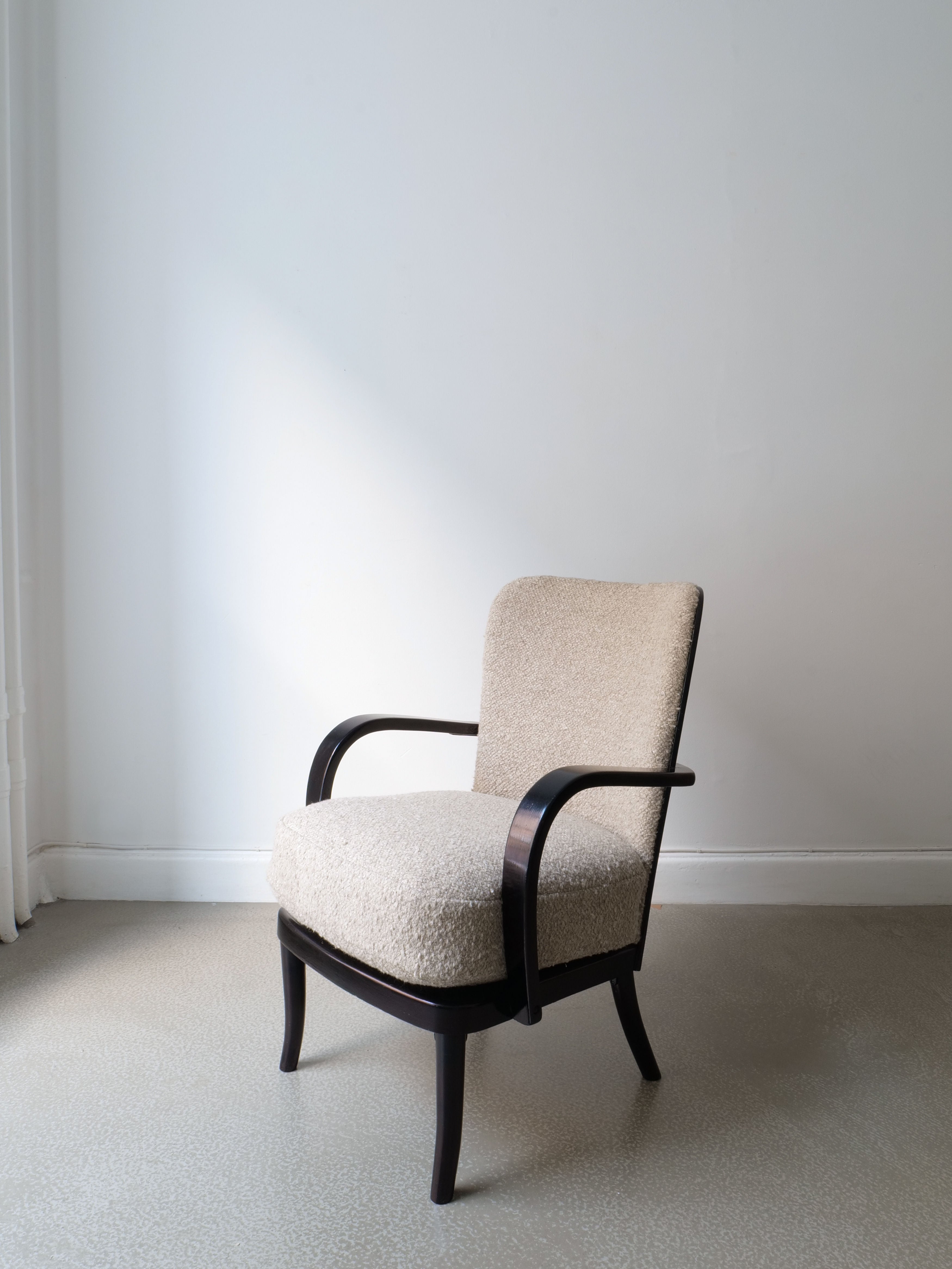 A pair of Werner West armchairs from the 1930s, part of the Collection apart brand, features a beige upholstered design with dark wooden arms and legs. These chairs rest on a light-colored floor against a plain white wall. The room's minimal decoration highlights the simple and elegant design of these classic armchairs.