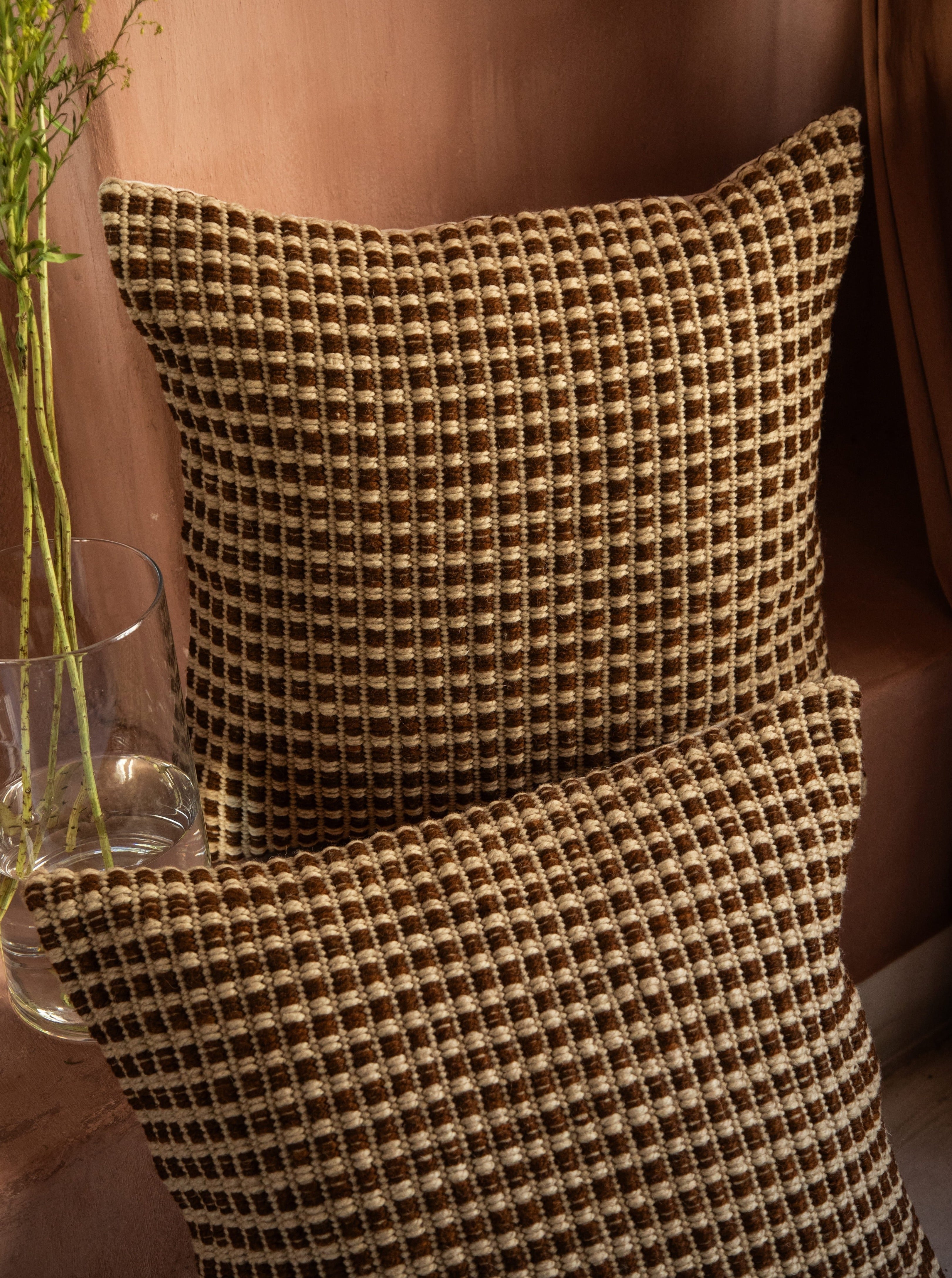 A textured brown and beige checkered Cima Pillow, crafted from hand-spun sheep wool by Identidad Argentina, rests against an elegant, smooth pink backdrop, with a clear glass vase containing sprigs of greenery beside it.