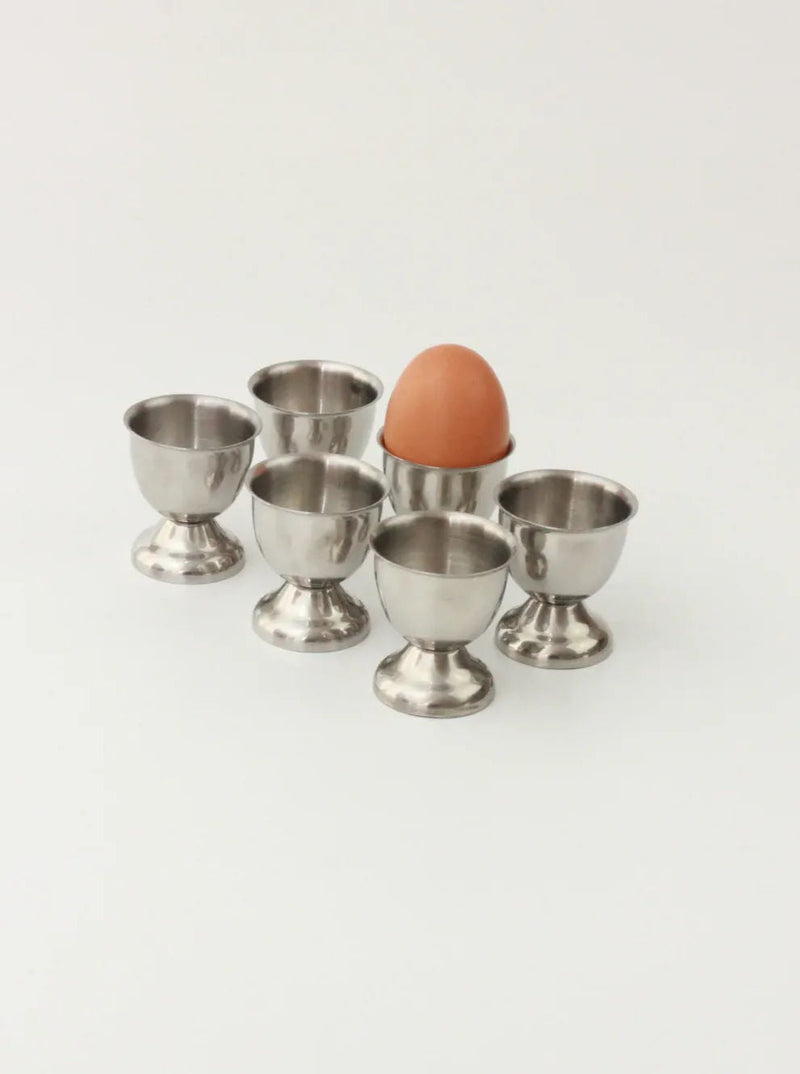Six durable stainless steel egg cups from the Set of Six Stainless Steel Egg Cups by Boga Avante Shop are arranged in a group on a plain white surface. One of the elegant breakfast tableware cups holds a brown egg, while the other five remain empty.