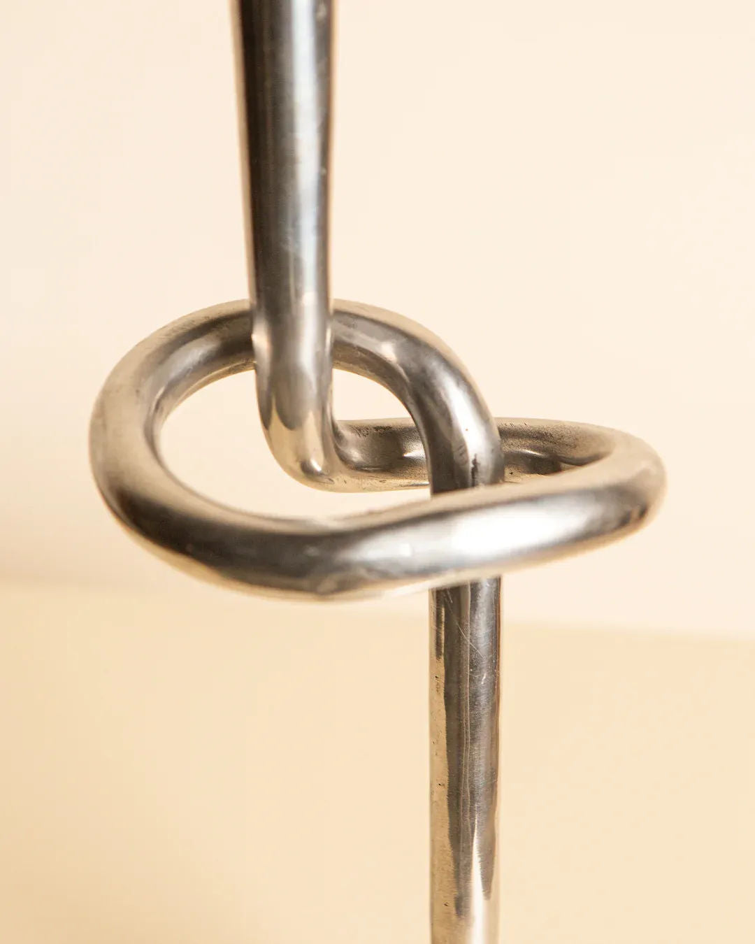 An intricate close-up of the Treaptyque Aluminum “Nœud” Candleholder 80's displays its sculptural design, with a looped knot in the middle set against a neutral background. The polished aluminum surface reflects light beautifully, underscoring the modern elegance of its twists and curves.