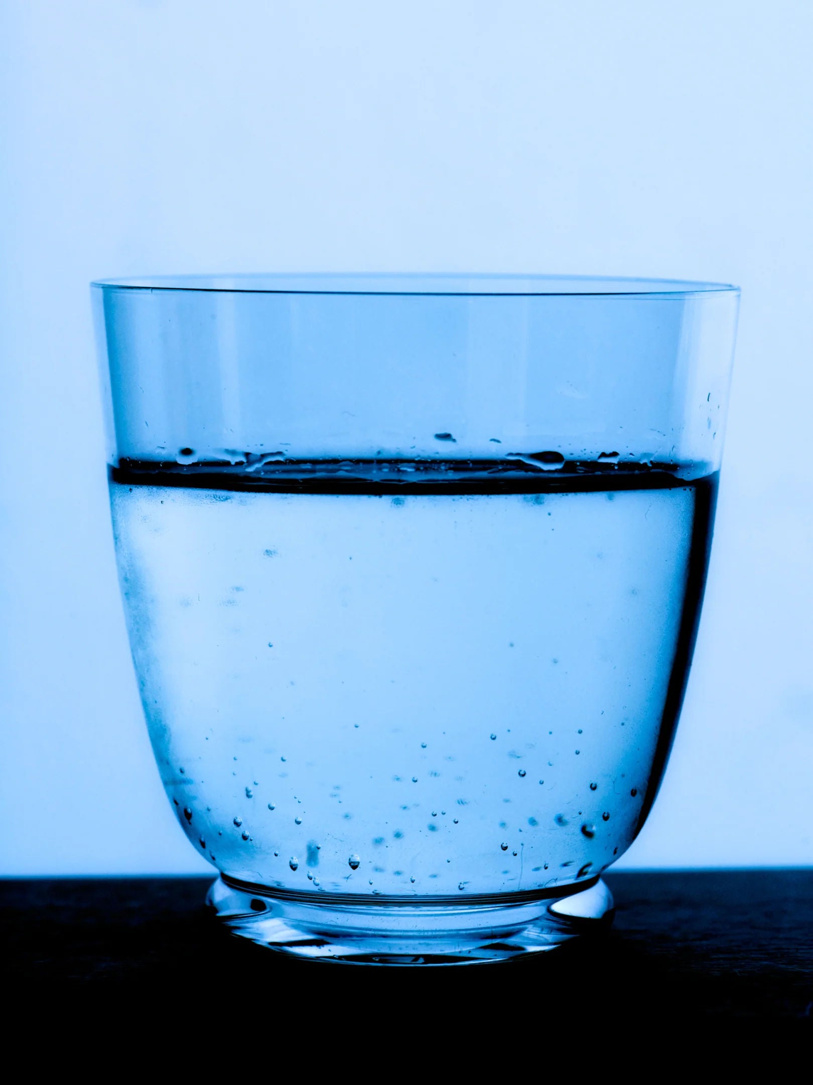 A Walter 01 Tumbler from the Service Projects collection, filled halfway with water and adorned with small bubbles, is set against a blue background. The simplicity and minimalistic design showcase the purity and transparency of the lead-free crystal.