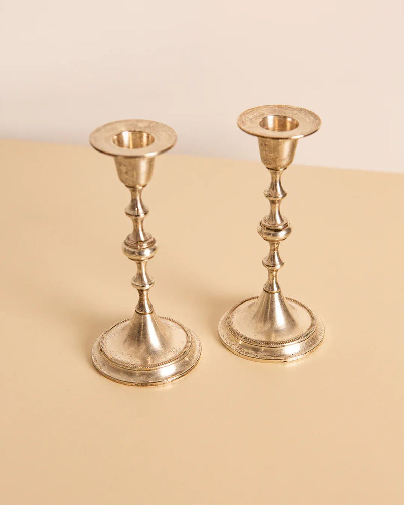 Two sophisticated silver candleholders, inspired by 1960s design, from Treaptyque adorn a light beige surface. These candleholders feature a timeless design with a flared base, slender stem, and round holders for candles.