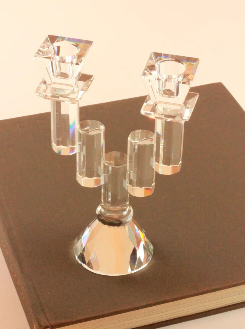 A Faceted Crystal Candleholder from Boga Avante Shop with three candle holders sits on a closed, dark brown book. The candleholder boasts a reflective, faceted surface reminiscent of an optical glass chandelier, catching the light to create a prismatic effect. This vintage item stands out against the plain, light-colored background.