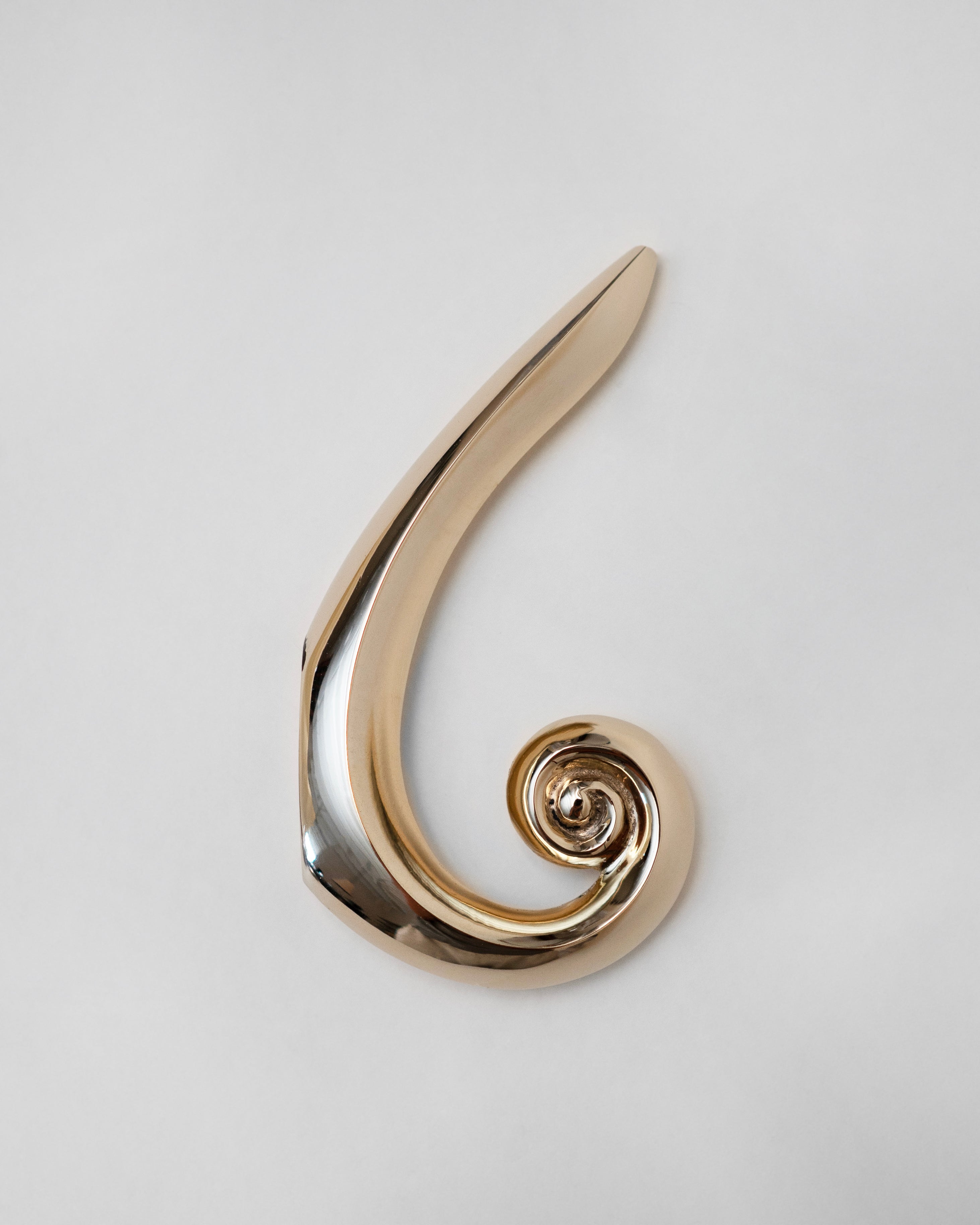 The Noelle Hook - Brass by Hein Studio features a shiny, metallic gold, abstract spiral design reminiscent of sandcasted hooks, standing out against a plain light gray background.