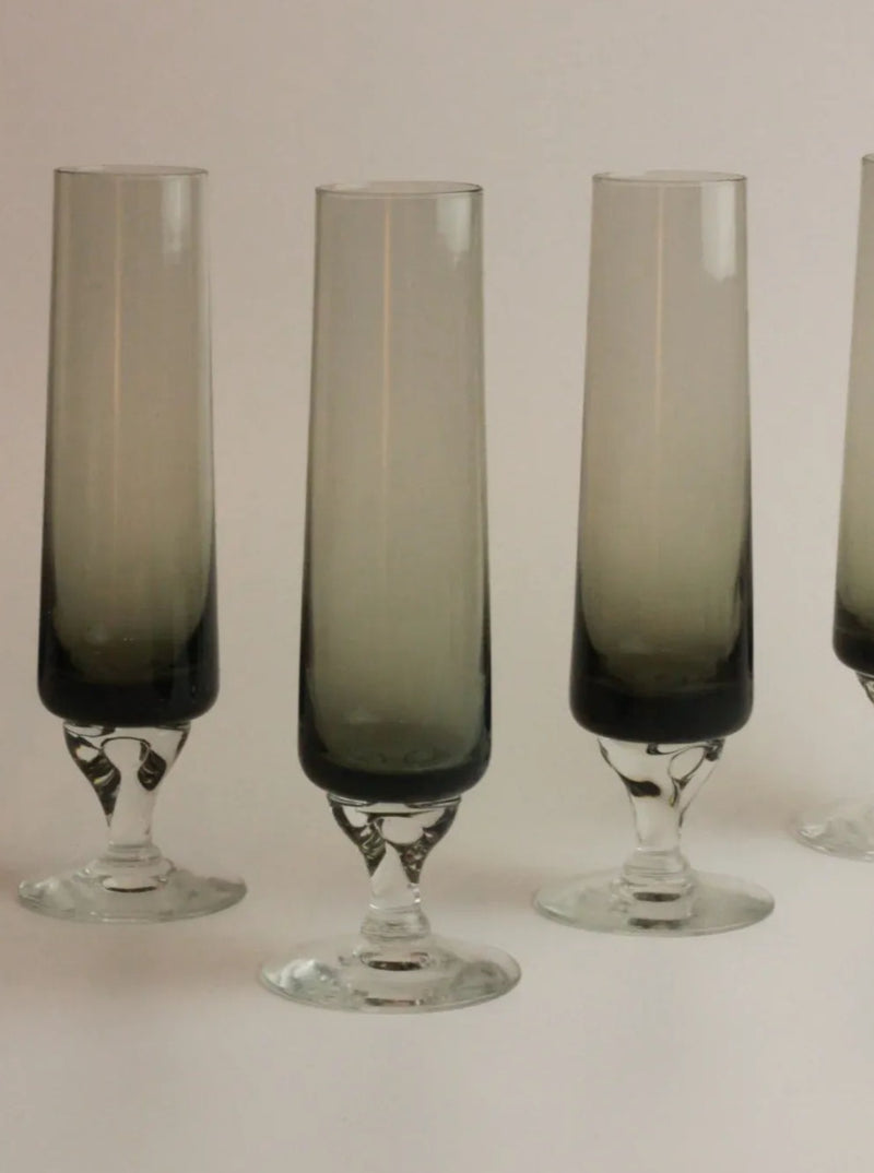 Four tall, slender, smoky grey champagne flutes with vintage flute-shaped design are arranged side by side on a neutral background. The flutes have a gradient design, becoming darker towards the base. This is the Set of Five Vintage Flute by Boga Avante Shop.