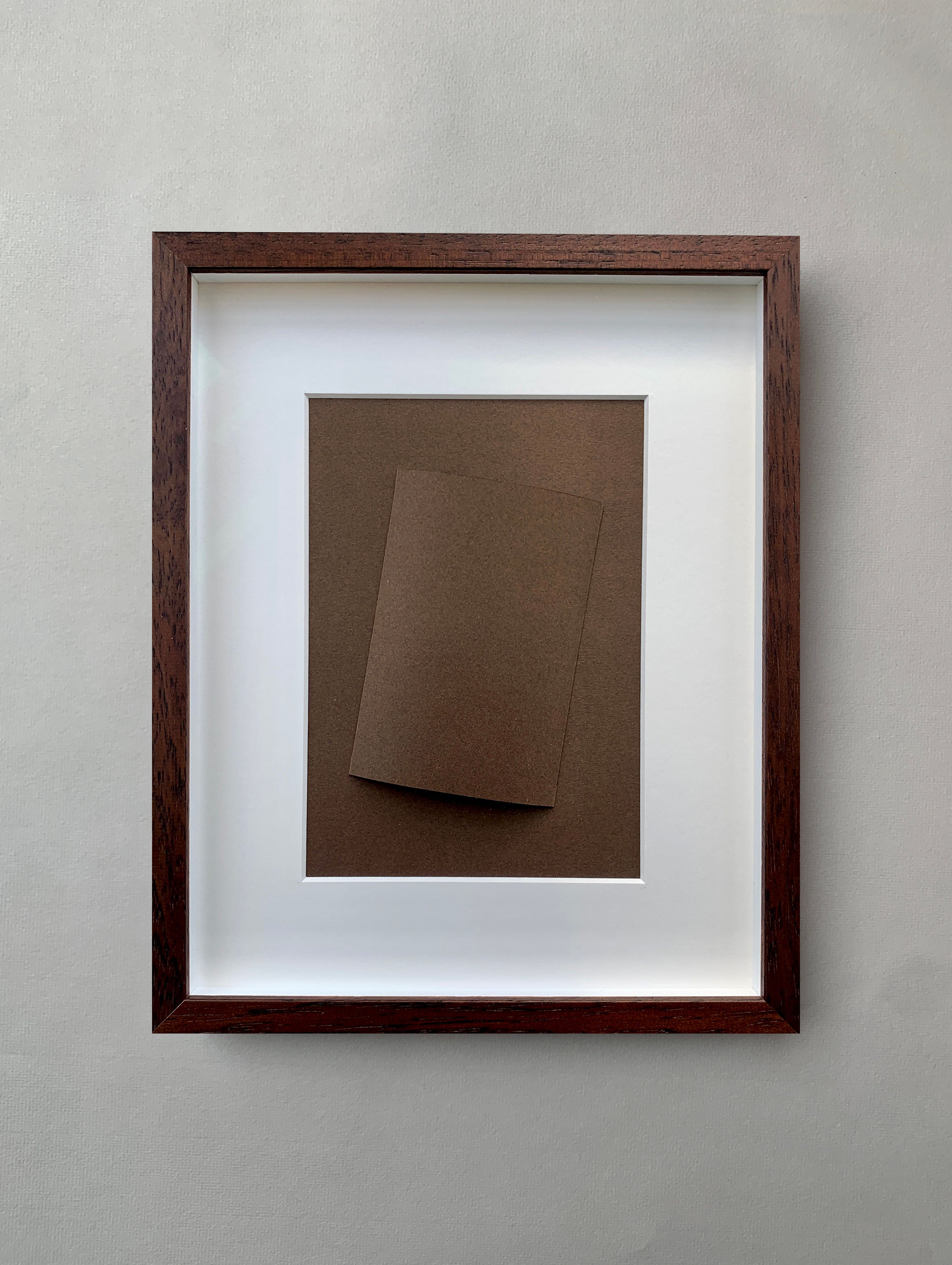 Marie Pomme Adriaansen's "Cylinder Brown - Paper Fold" features a framed abstract art piece with a monochromatic brown rectangular paper, slightly tilted to suggest architectural forms. It is set against a plain gray wall, encased in a wooden frame with a white mat.