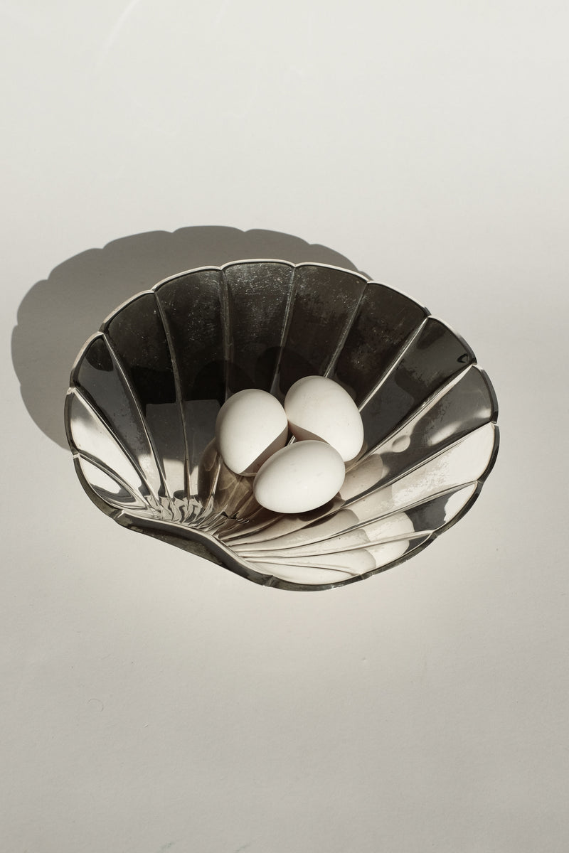 A Large Silverplated Shell Dish by septembre studios, featuring a wavy design, elegantly holds three white eggs. The decorative bowl rests on a light surface, casting a gentle shadow.