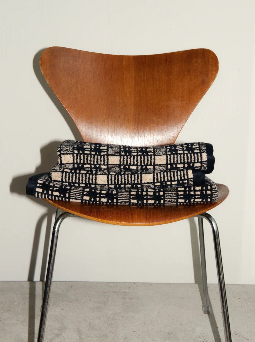 Modern wooden chair with a butterfly-shaped backrest and shiny chrome legs, featuring a black and white checkered cushion placed on the seat, set against a neutral wall with a shadow cast behind.