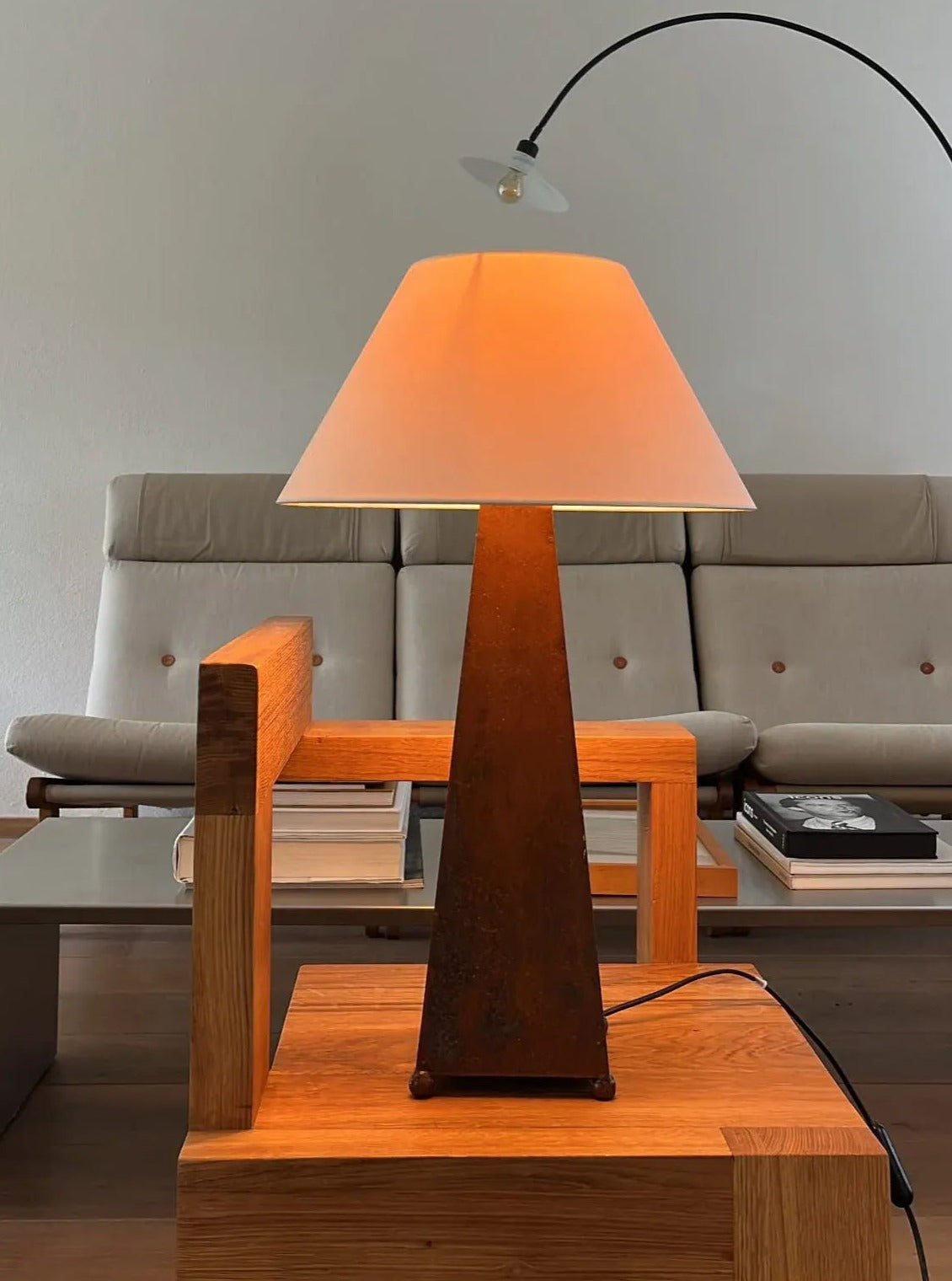 A modern, minimalist living room features a geometric wooden side table with the stylish CU-RA-TED Obelisk Table Lamp, showcasing a rust-colored, pyramid-shaped base topped with a beige lampshade. A beige sofa with button-tufted cushions and stacked books on a low table are visible in the background.