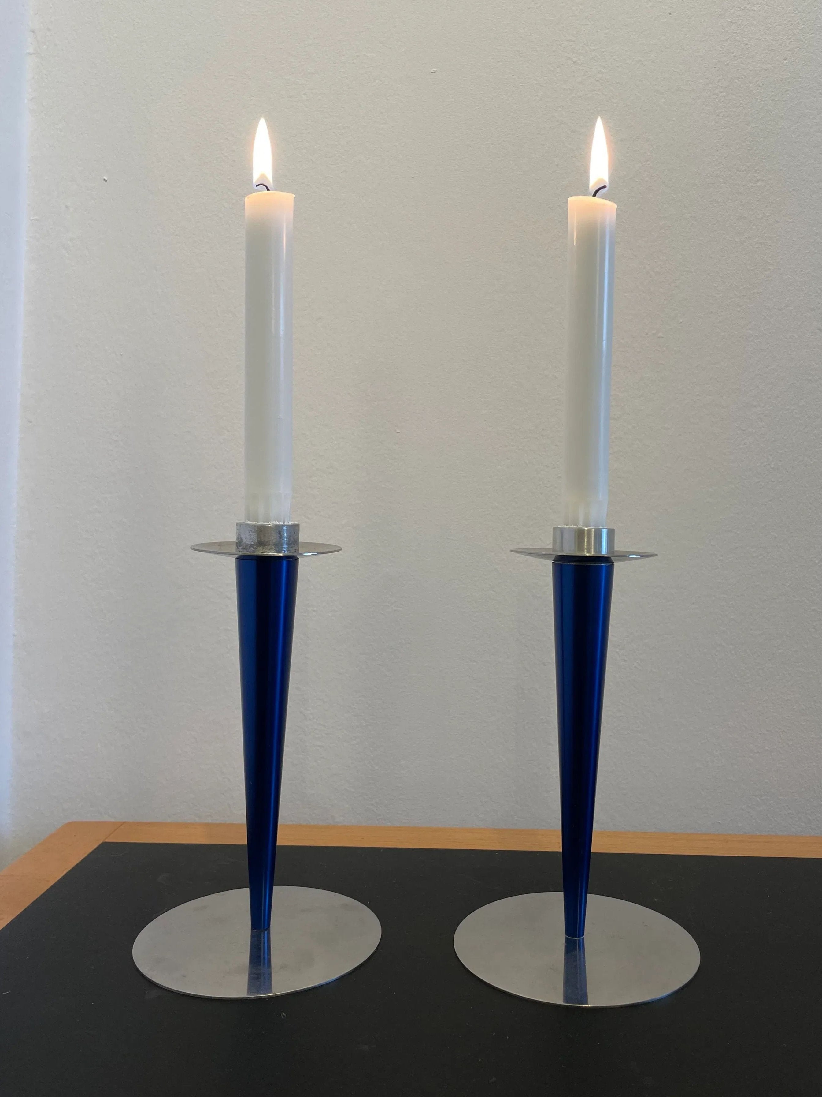 Two lit white candles are placed in sleek, modern blue and silver Vintage Steel Candle Holders by Collect Cph. The conical holders, reminiscent of a vintage menu design from Denmark, taper elegantly from a wide base to a narrow top. They rest on a dark surface against a plain, light-colored wall.