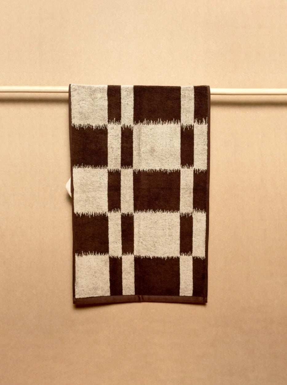 An Autumn Sonata Karin Hand Towel with a brown and white checkered pattern hangs neatly on a horizontal bar against a plain beige background.