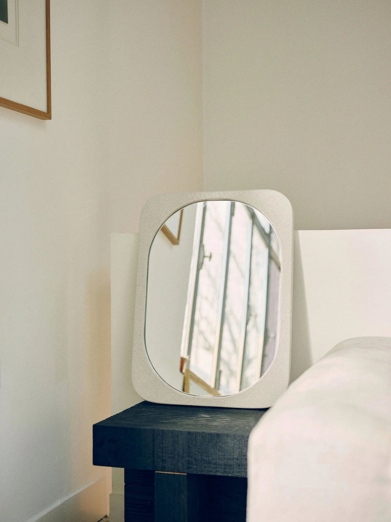 A modern, rectangular artisanal mirror with rounded edges and a crackled enamel finish, the Mirror Bien Fait in Lava Stone by Bien Paris, is propped up on a dark wooden side table against a white wall. The mirror reflects a staircase with thin vertical railings, while part of white framed artwork and a light-colored couch are visible in the background.