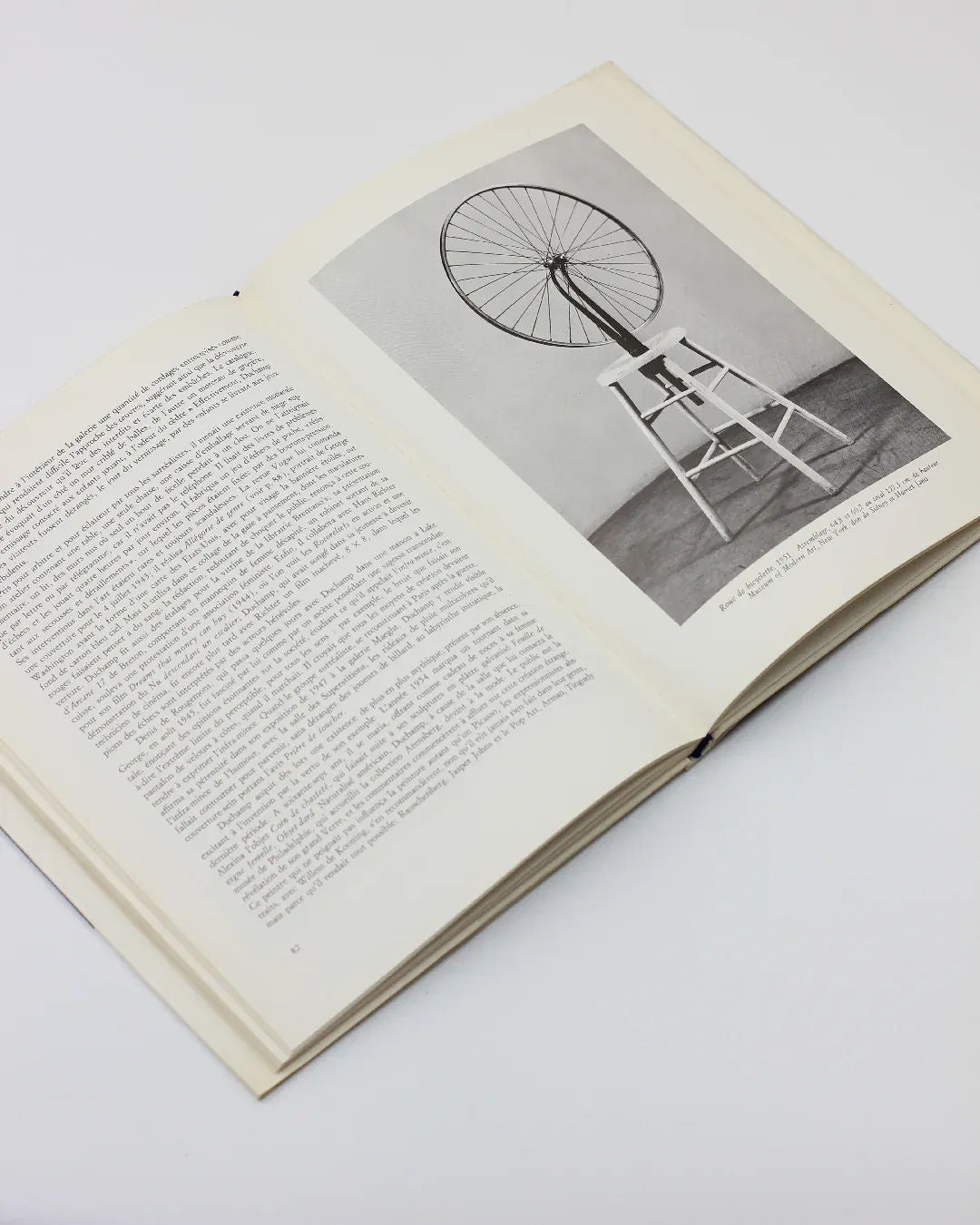 An open copy of "Marcel Duchamp by Sarane Alexandrian Book" from Boga Avante Shop reveals dense text on the left page, while the right page features a black-and-white photograph of a bicycle wheel mounted on a white stool. This minimalist composition reflects Marcel Duchamp's conceptual art, where ordinary objects are transformed into thought-provoking creations.