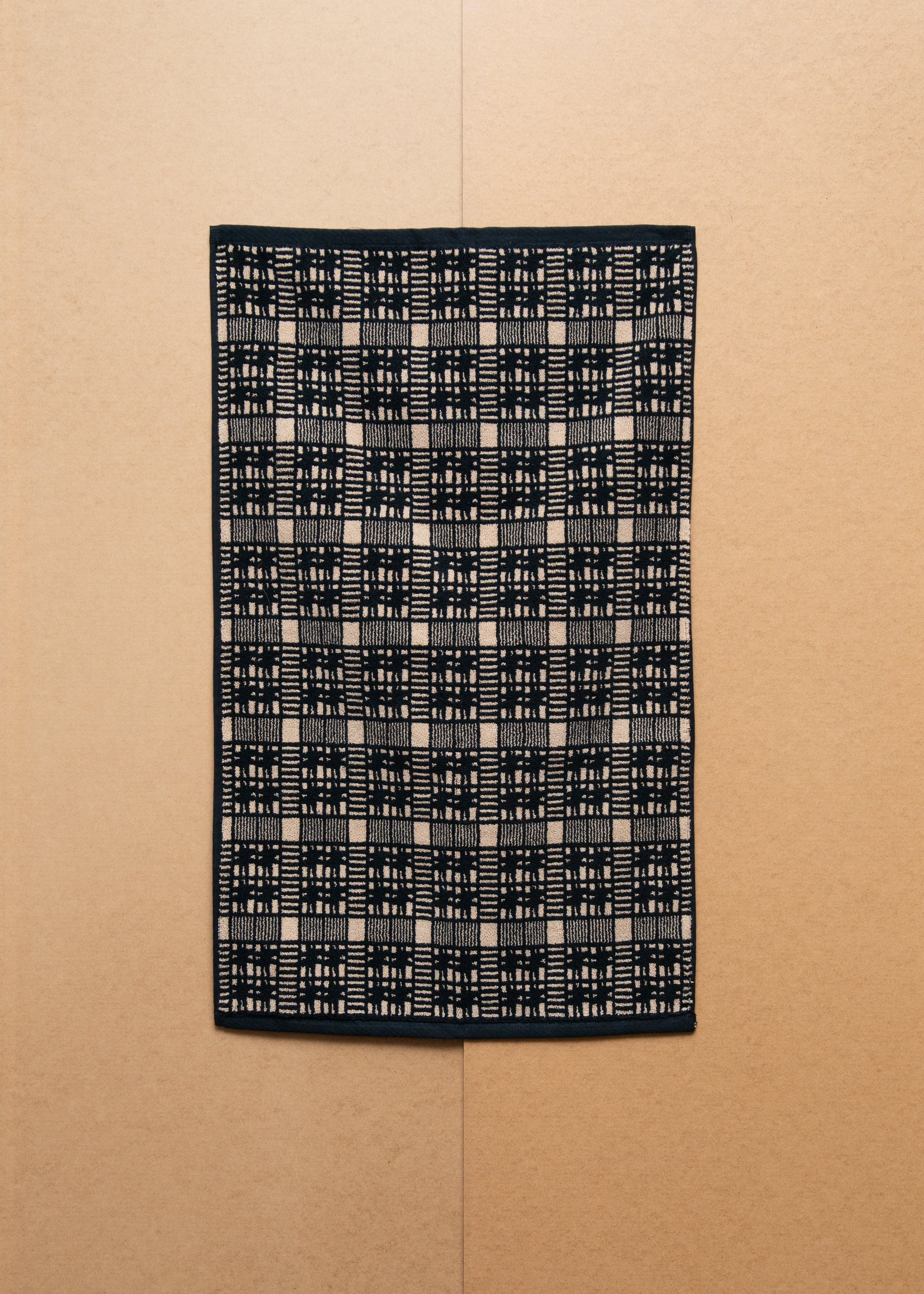 A rectangular black Alma Bath Mat towel with a white checkered design hung on a beige wall.