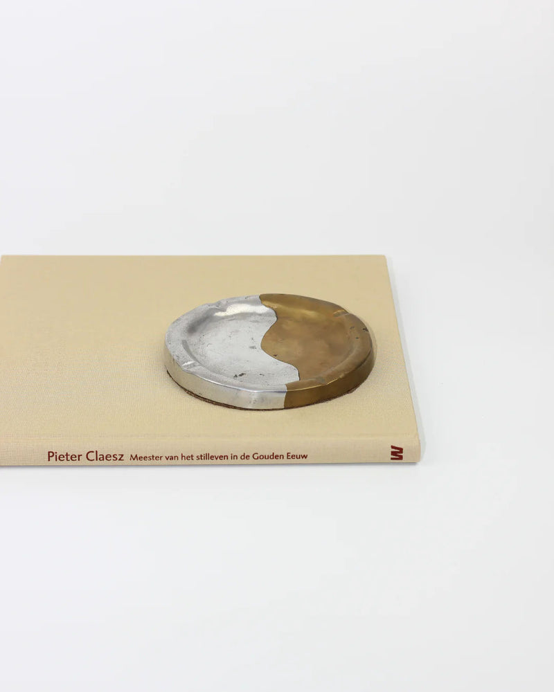 On a white surface rests the closed book "Pieter Claesz Meester van het stilleven in de Gouden Eeuw," topped by Boga Avante Shop's Brutalist Ashtray, a round metal object with half aluminum and half brass design.