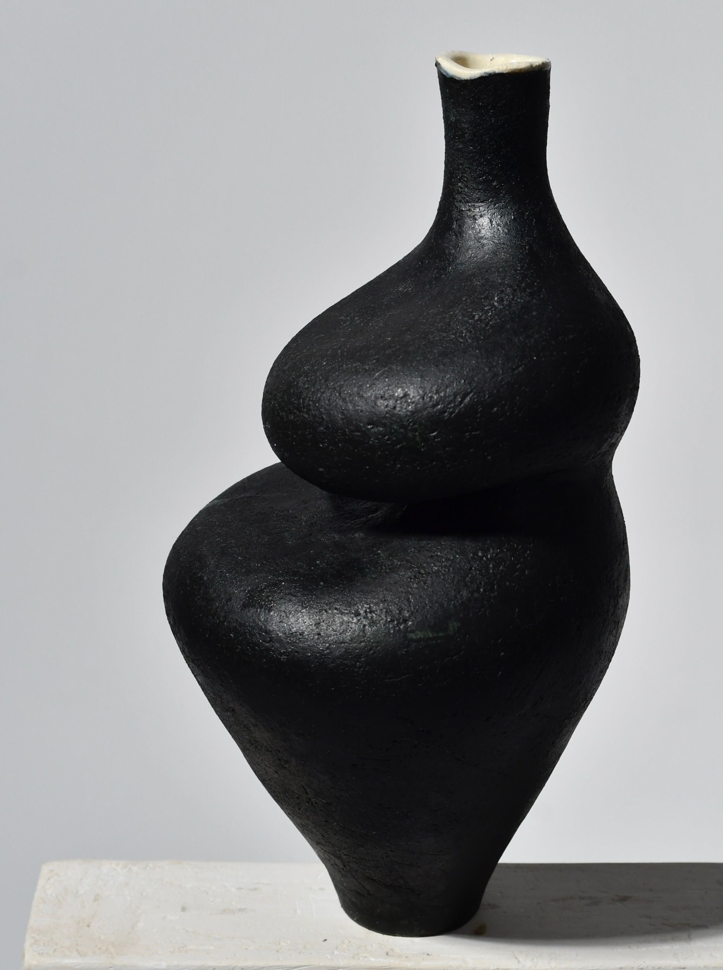 A modern black sculpture resembling a stacked, abstract form with a smooth, matte finish, displayed on a light-colored base against a neutral background. This sculptural vessel features intricate details and is positioned prominently to Black Lilith with Oxide Drip by Lucia Mondadori.