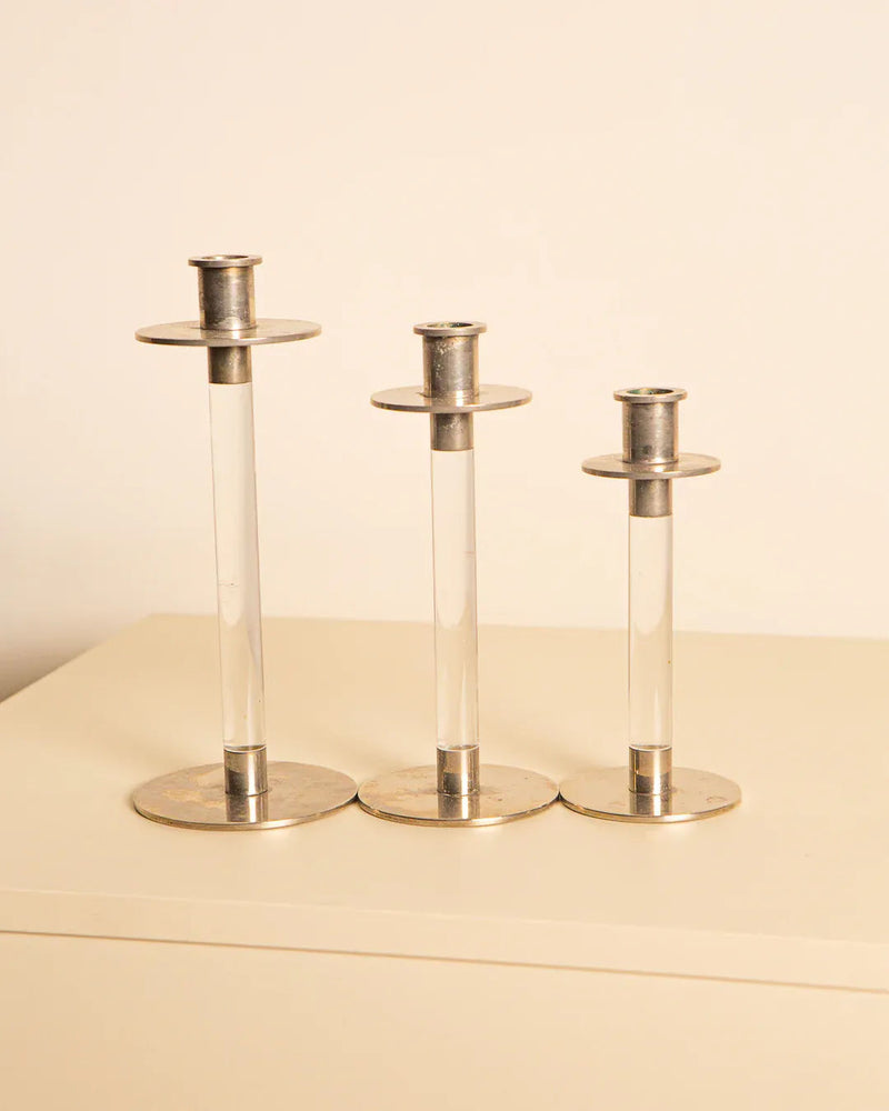 The Treaptyque Set of 3 80's Candleholders features clear cylindrical stems and sleek metallic bases, perfectly blending modern and retro styles. Displayed on a beige surface against a light cream background, they make ideal lucite silver accents for any space.