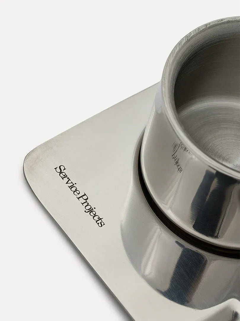 A stainless steel cup from The Arlo Espresso set by Service Projects rests on a square metal tray with "Service Projects" engraved on the edge. The Italian design's polished surfaces reflect light and nearby objects.