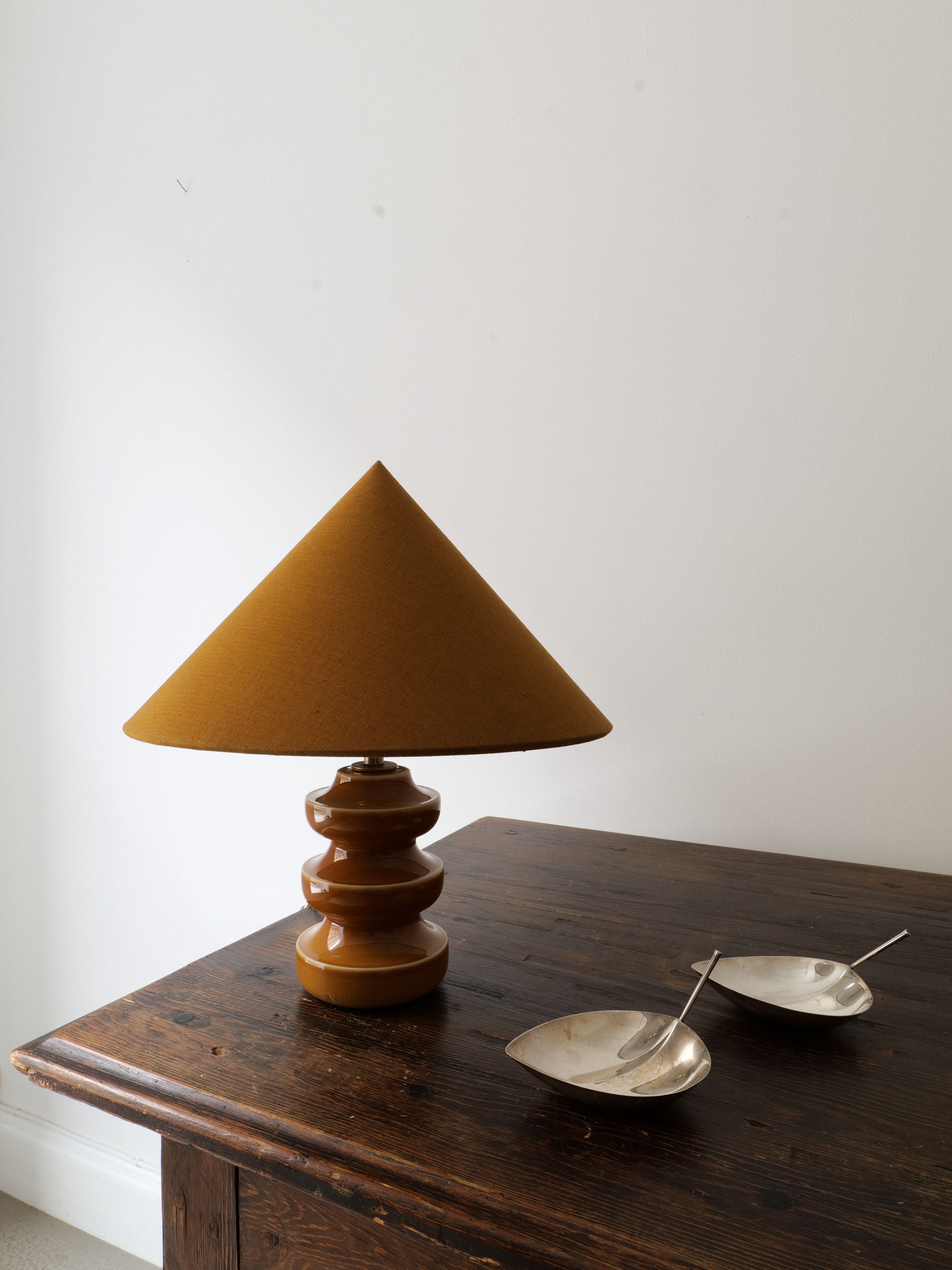 A side table holds a Small Sculptural Lamp by Collection apart, featuring an earthy-toned ceramic with a stacked, rounded base and a wide, conical shade. Next to the lamp are two silver, leaf-shaped dishes with small spoons. The background wall is plain white.