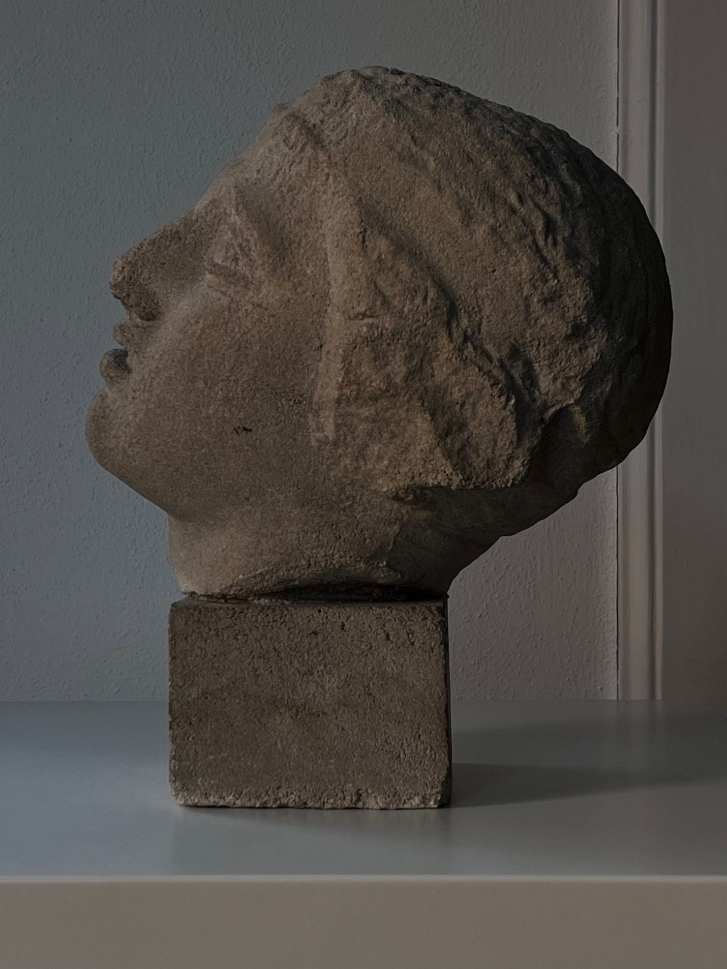 The Decorative Bust by Médecine, featuring a sandstone composite design with textured hair and a serene expression, rests on a square base. This mid-20th-century sculpture is elegantly placed on a smooth surface against a plain wall, with side lighting creating gentle shadows.