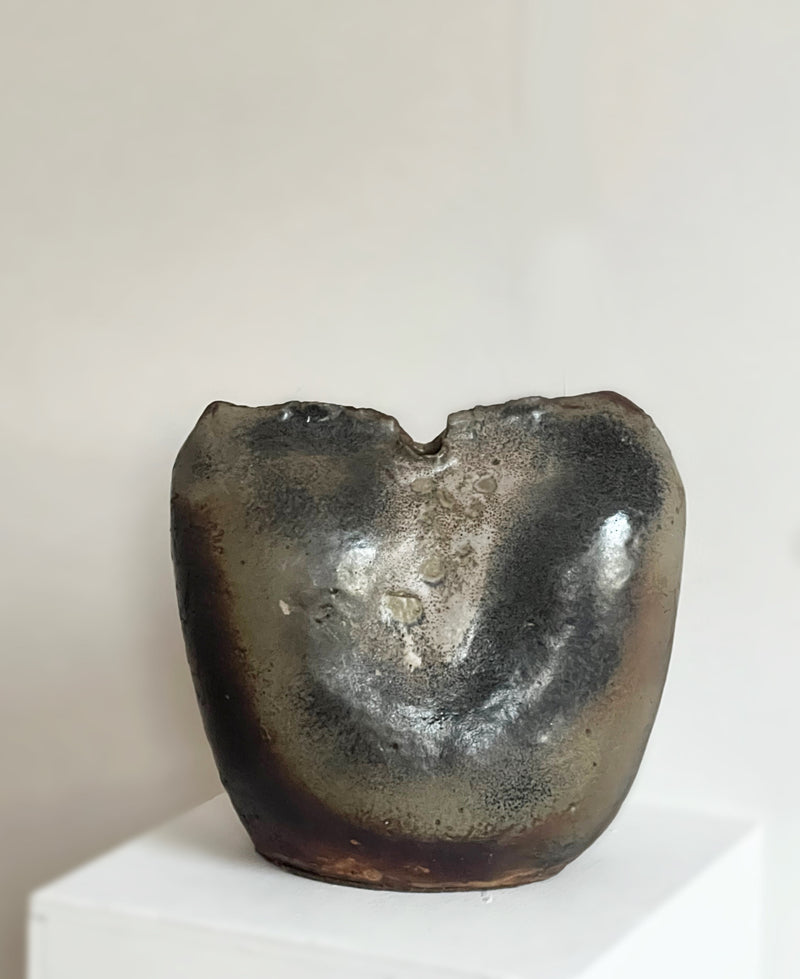 The Porter Studio Ceramic Pinch Top Studio Vase, featuring a metallic texture, abstract organic form, and an uneven split top edge, sits on a white pedestal against a plain light background.