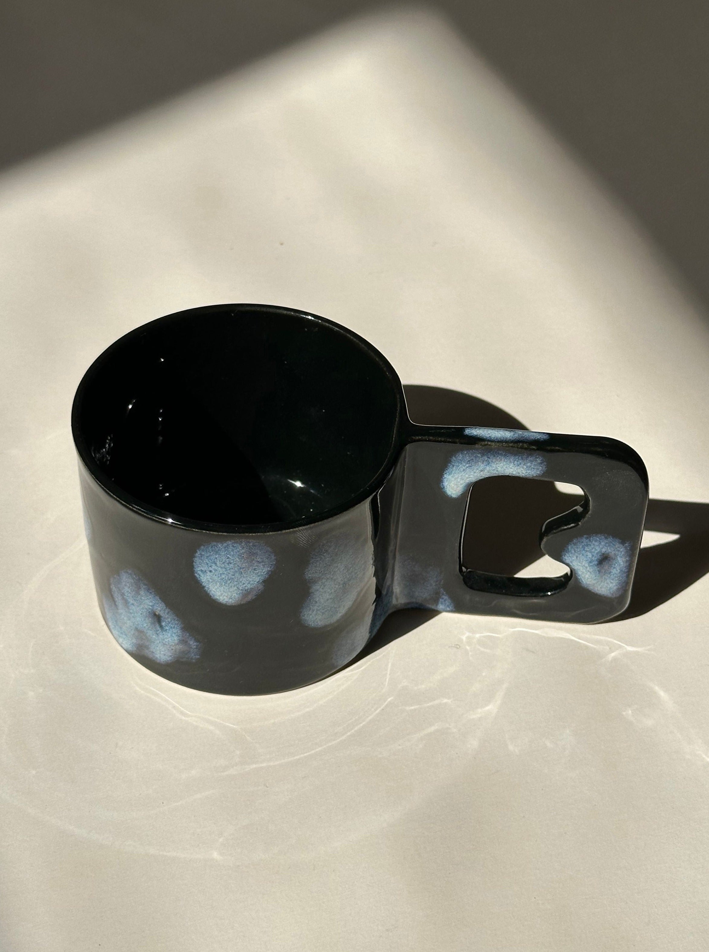 A ceramic mug crafted from stoneware clay, featuring a black glaze with irregular light blue spots, rests on a light-colored surface. The angular handle, complete with a heart-shaped cutout, is perfectly positioned to showcase noki ceramics' Cloud Cup charm and its beautifully glazed interior.