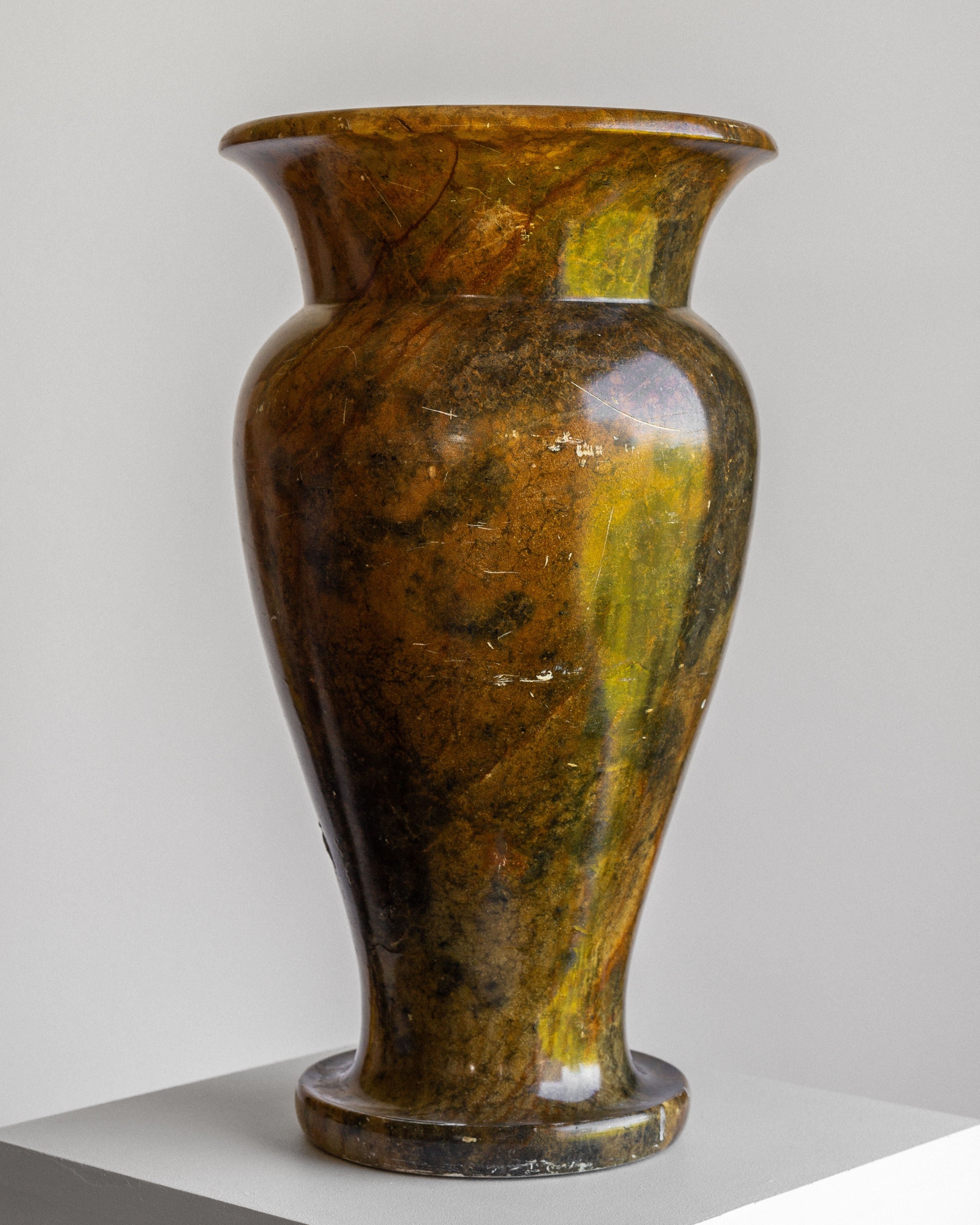 An elegant, glossy Large Neoclassical Stone Vase by Spigel, with a flared rim and wide-bodied, urn-shaped design, featuring a rich marbled pattern in shades of brown, yellow, and green. The vase is positioned on a white pedestal against a light gray background, highlighting its earthy tones and smooth finish.