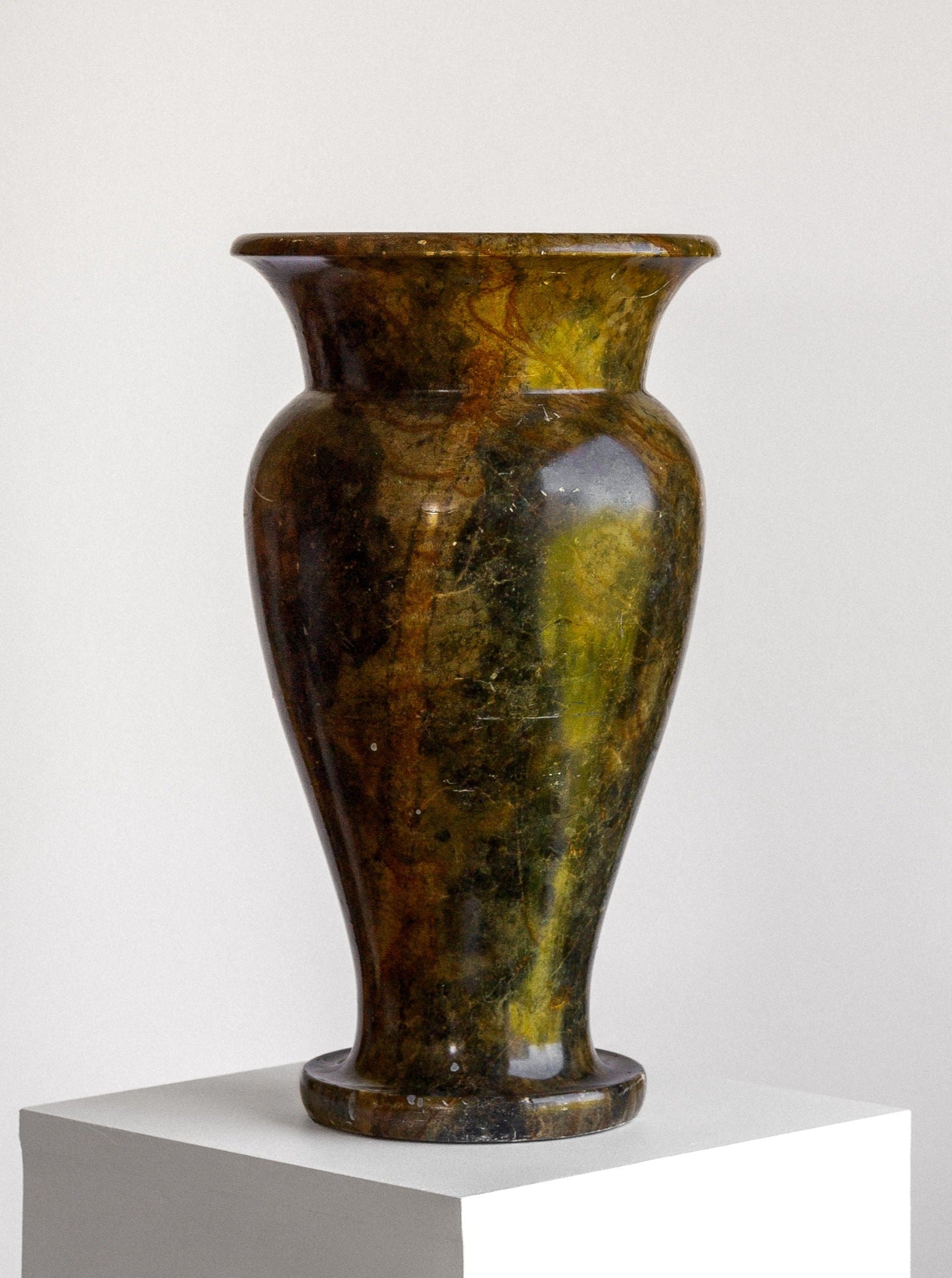 A Large Neoclassical Stone Vase by Spigel, featuring a tall urn-shaped design with an elegant classical shape, sits on a white pedestal. Its glossy marbled green and brown surface showcases an intricate pattern and shine against the plain white background. The wide rim and narrow base highlight its polished details.