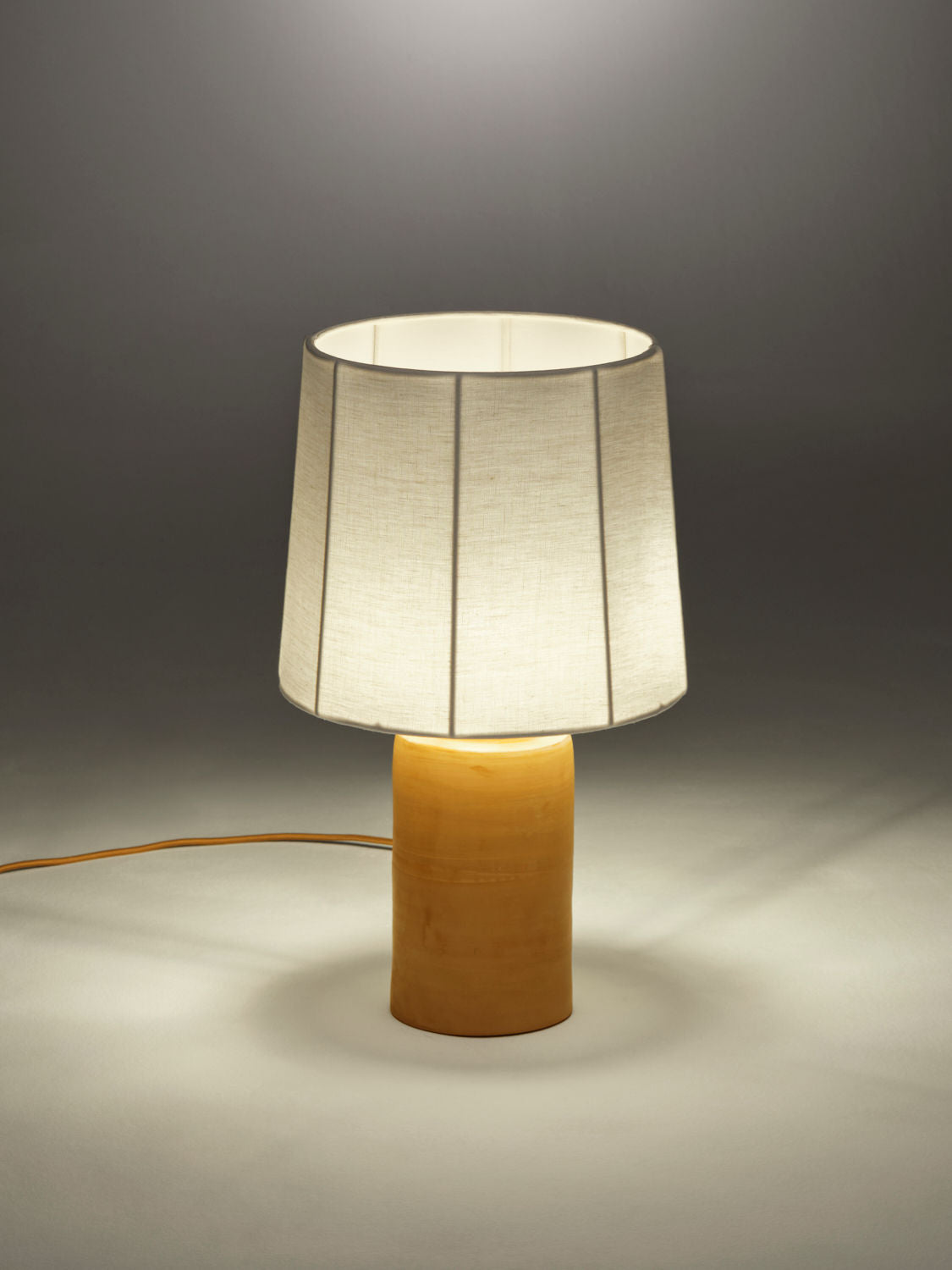 A Nassi Lamps Sonora Linen Table Lamp with a wooden cylindrical base topped by a wide, pure linen lampshade. The lamp is illuminated, casting a soft glow around it. A power cord is visible extending from the base. The background is a plain, neutral color.