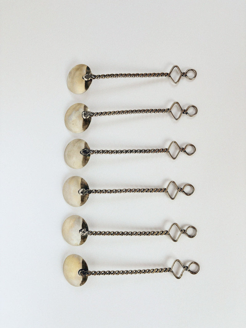 Six identical items, like functional art objects, are aligned vertically on a white surface. Each features a circular disk and a lobster clasp linked by a twisted chain segment, similar to the 70's Handmade Spoons Set by Huerto.